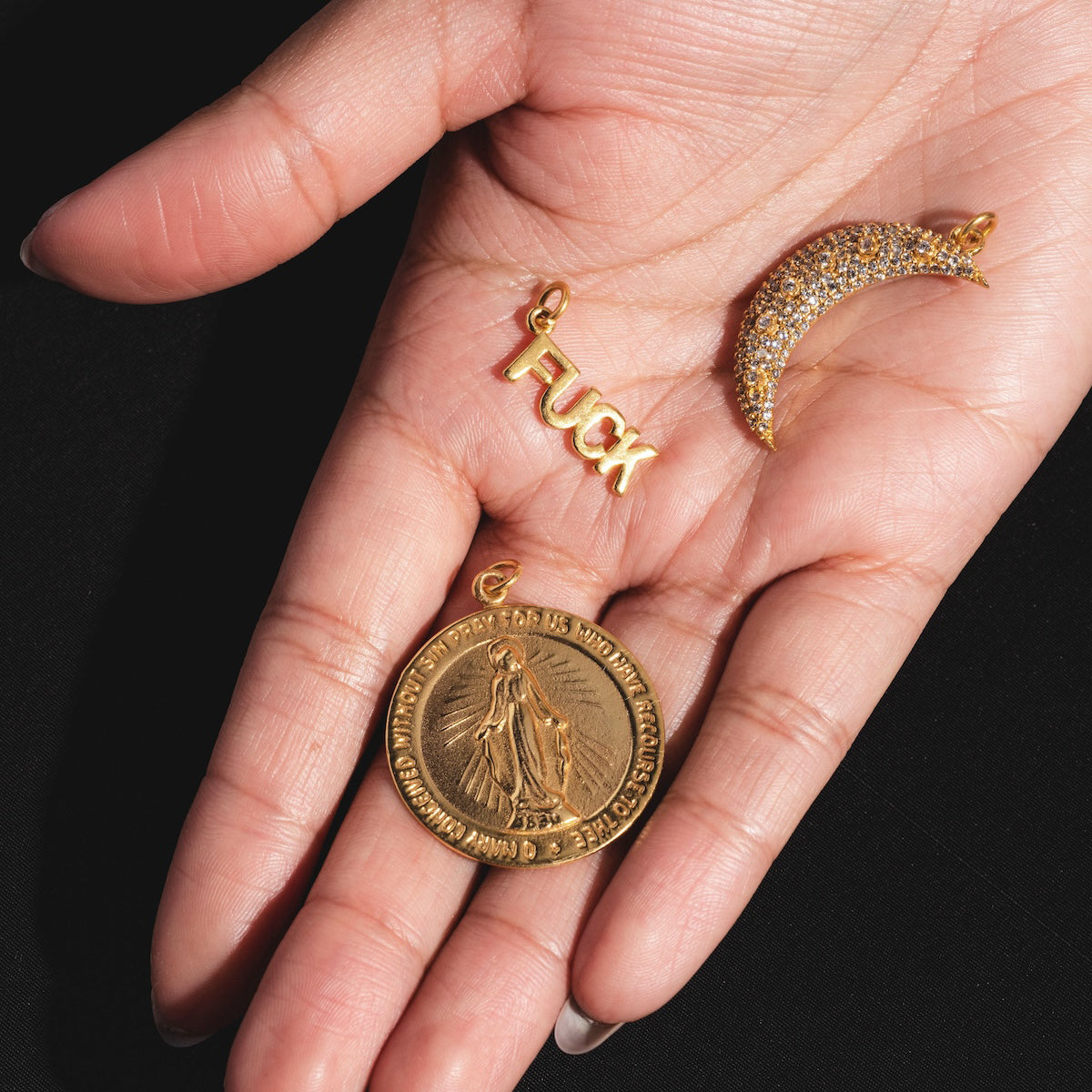 A hand holds three gold-plated Charms by Anokhi Green: one spelling Fuck, a crescent moon encrusted with green stones, and a round Virgin Mary medallion inscribed along the edge.