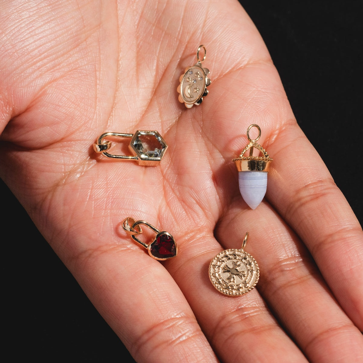 A hand displays five small charms—including a Large Blue Agate Amphora Charm, solid gold charm, red heart, hexagonal gem, and oval locket—set beautifully against a black background.