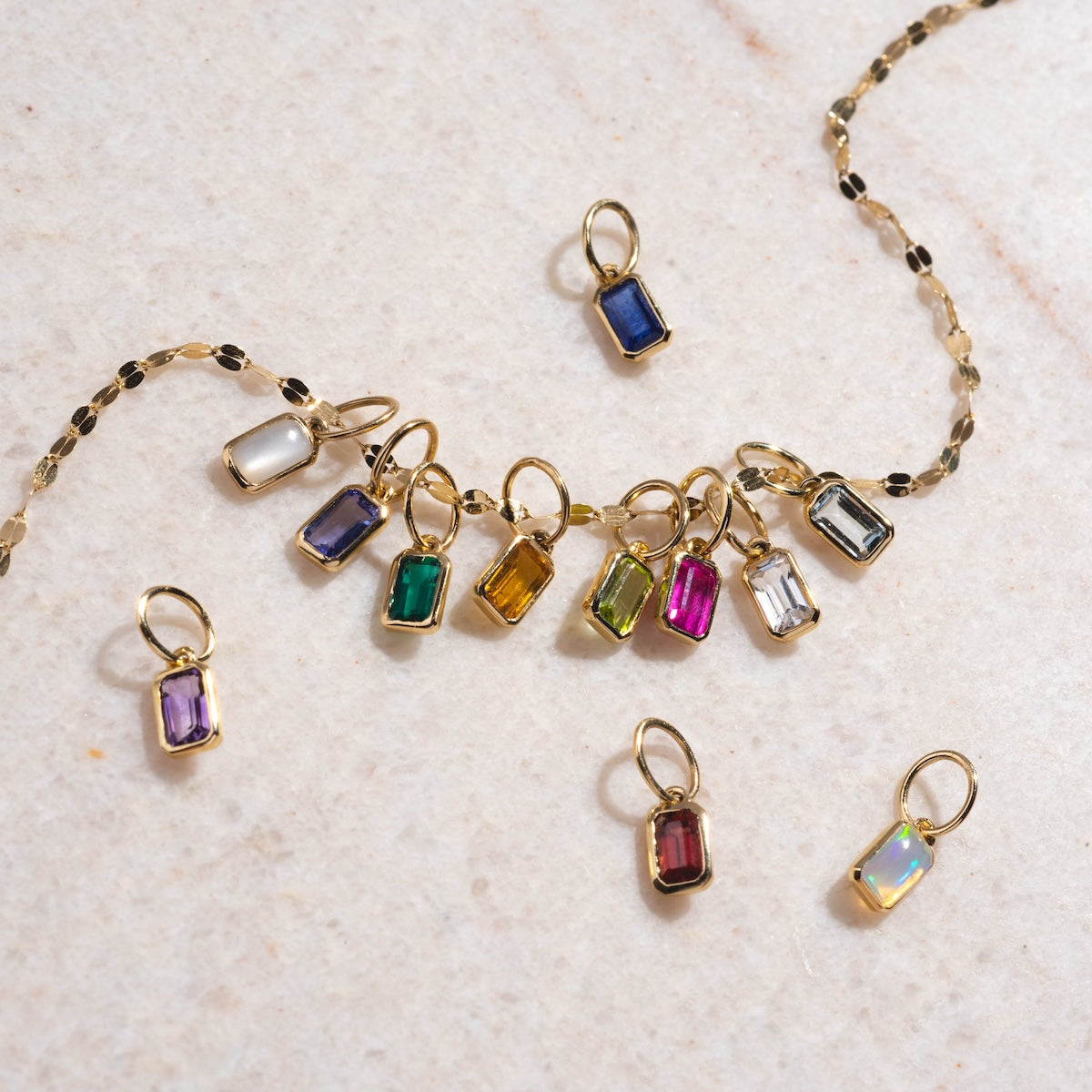 A gold chain features twelve Solid Gold Birthstone Baguette Charms in vibrant colors, each set in a gold frame and displayed on light marble—an ideal personalized gift for jewelry lovers.