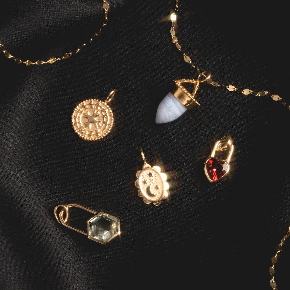 Five gold charms and pendants—including a Large Blue Agate Amphora Charm, a coin, red heart, green hexagon, and crescent moon round charm—are displayed on black fabric beside a solid gold chain necklace.
