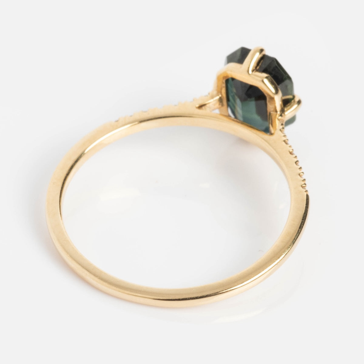The Fira Ring with Sapphire features a faceted, dark green hexagonal sapphire in a prong-set solid gold band, shown from the side and slightly above on a white background—an elegant choice for teal gemstone lovers.