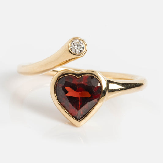 The Heart Garnet and Diamond Twist Ring is crafted in 14k solid gold, showcasing a heart-shaped red garnet and a small round diamond on an open, curved band against a white background.