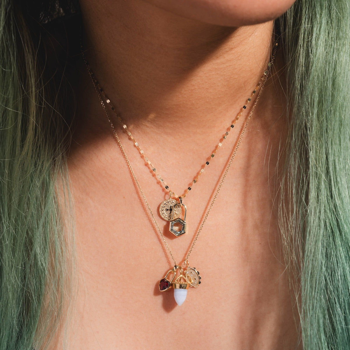 A close-up of a person’s neck with light green hair, wearing two layered gold necklaces. One necklace features a geometric pendant; the other showcases the Large Blue Agate Amphora Charm and a small white stone.