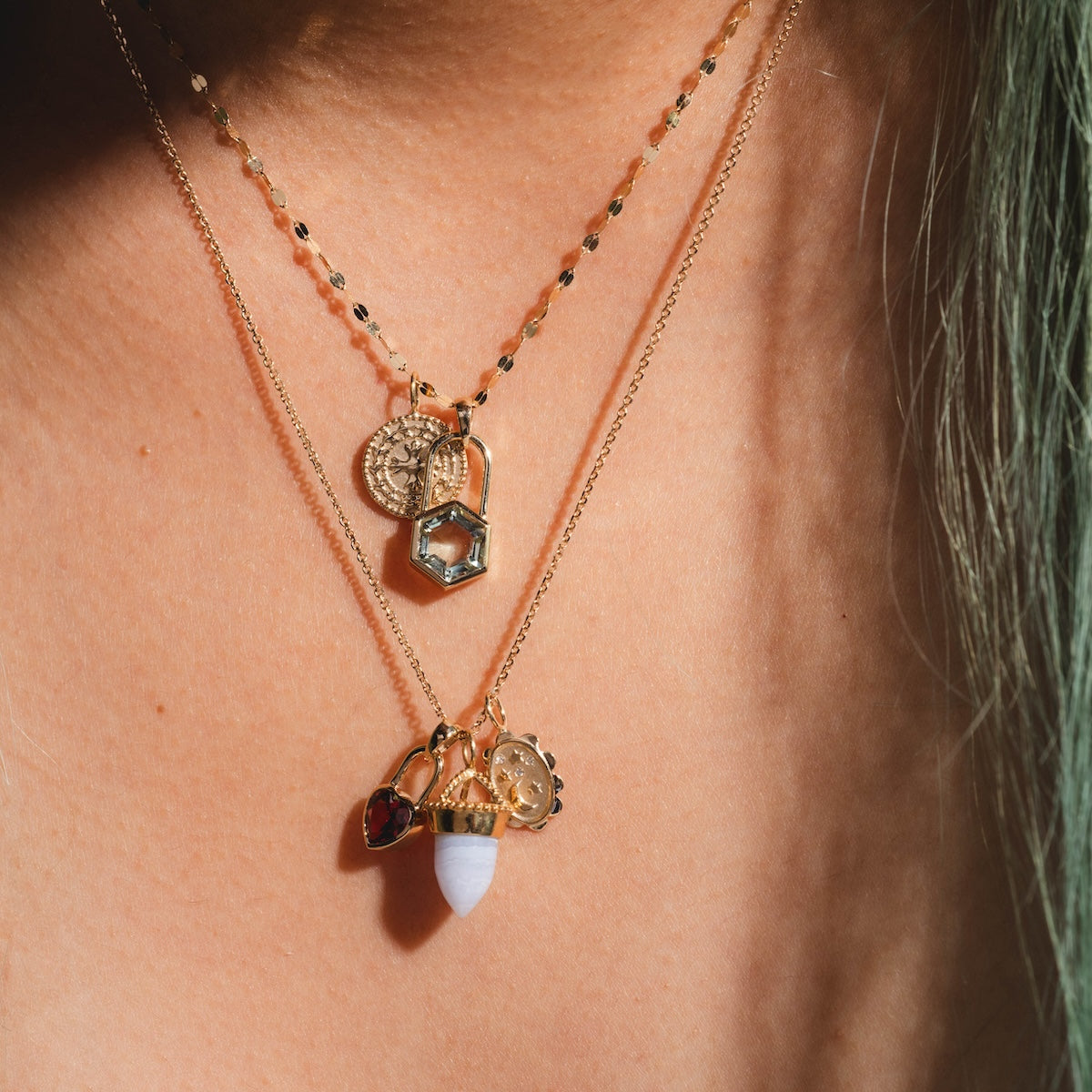 A close-up of a person’s neck wearing two gold necklaces featuring several charms, including a solid gold piece, a coin, a red heart, hexagon, white stone, and the Large Blue Agate Amphora Charm.