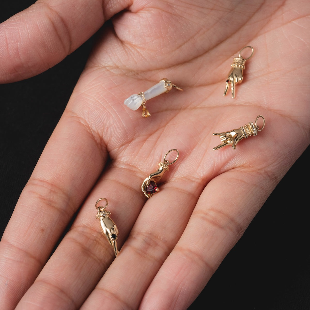 A hand holds five intricately detailed Solid Gold Body Language Charms, each with unique gestures. One exclusive Local Eclectic charm features a clear crystal, all set against a striking black background.