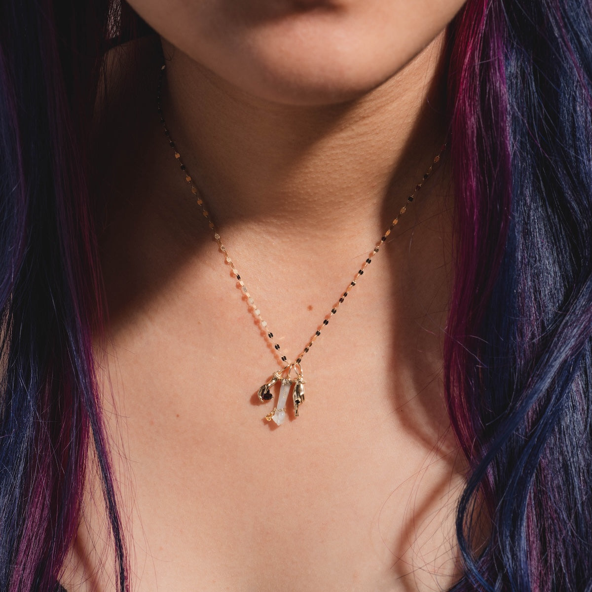 A close-up of a woman wearing the exclusive Local Eclectic Solid Gold Body Language Charm necklace, featuring alphabet and crystal pendants. Her straight, purple and blue hair frames her face, accentuating her effortless style.