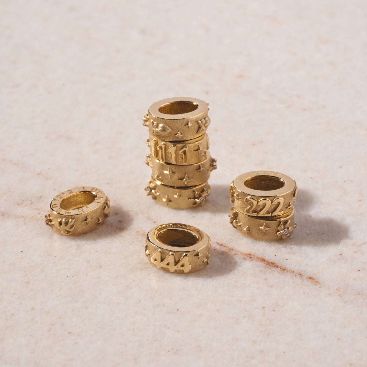 Five Solid Gold Angel Number Spacer Charms with engraved numbers (111, 222, 333, 444, 777) and star-shaped details, arranged on a light speckled surface. A Local Eclectic Exclusive—four stacked vertically, three separate.