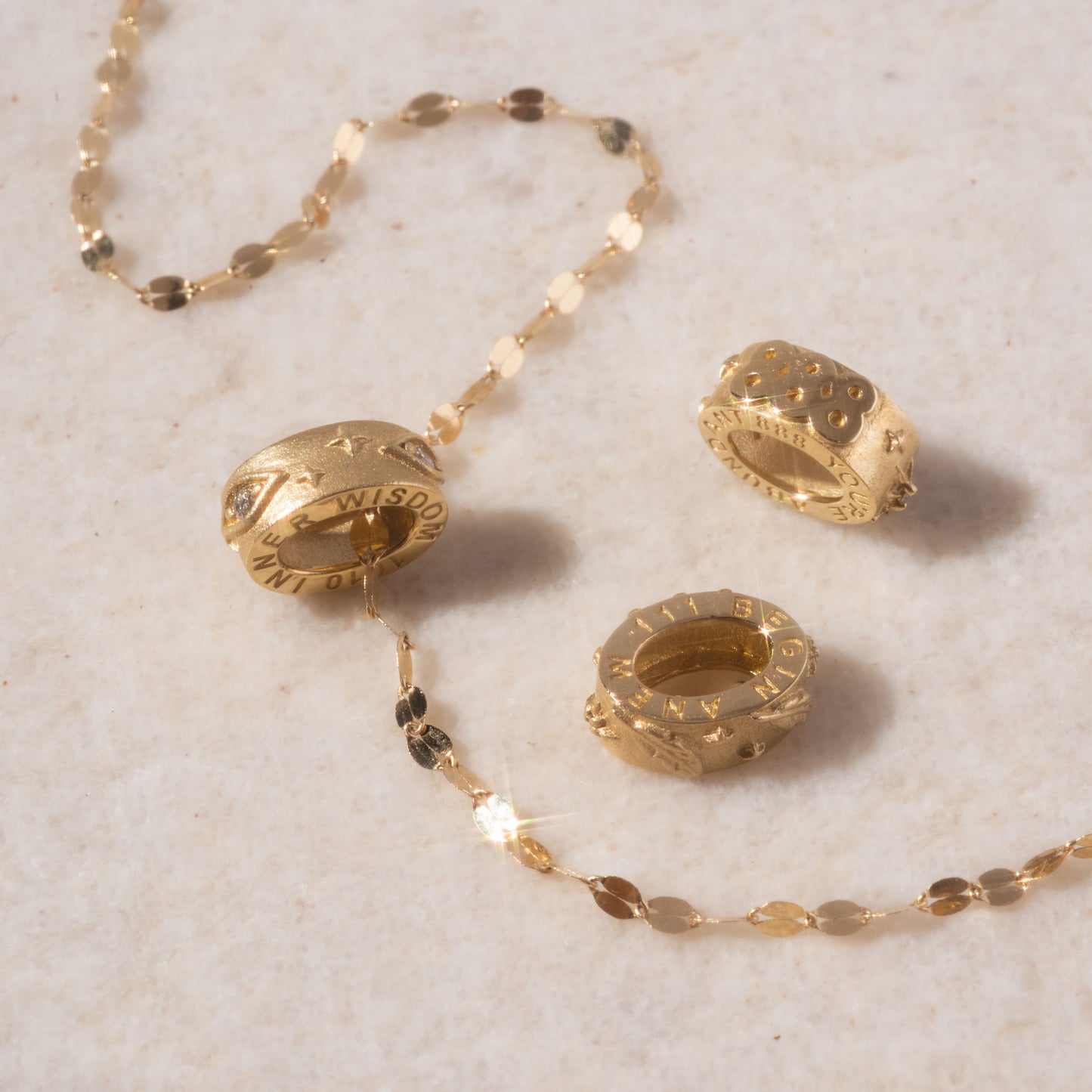Three textured gold beads with engraved numbers rest on a beige surface, one bead threaded onto an oval-link chain. The round, chunky Solid Gold Angel Number Spacer Charm is perfect for a Local Eclectic Exclusive look.
