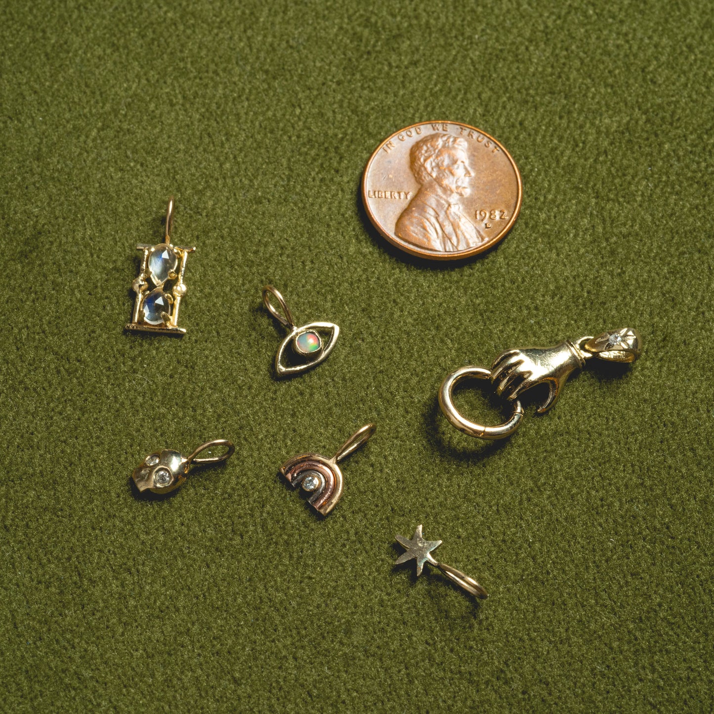 Seven 14k solid gold tiny charms by Acanthus—hourglass, eye, hand with ring, star, heart, and two abstract shapes—are shown on green fabric beside a 1983 U.S. penny for scale.