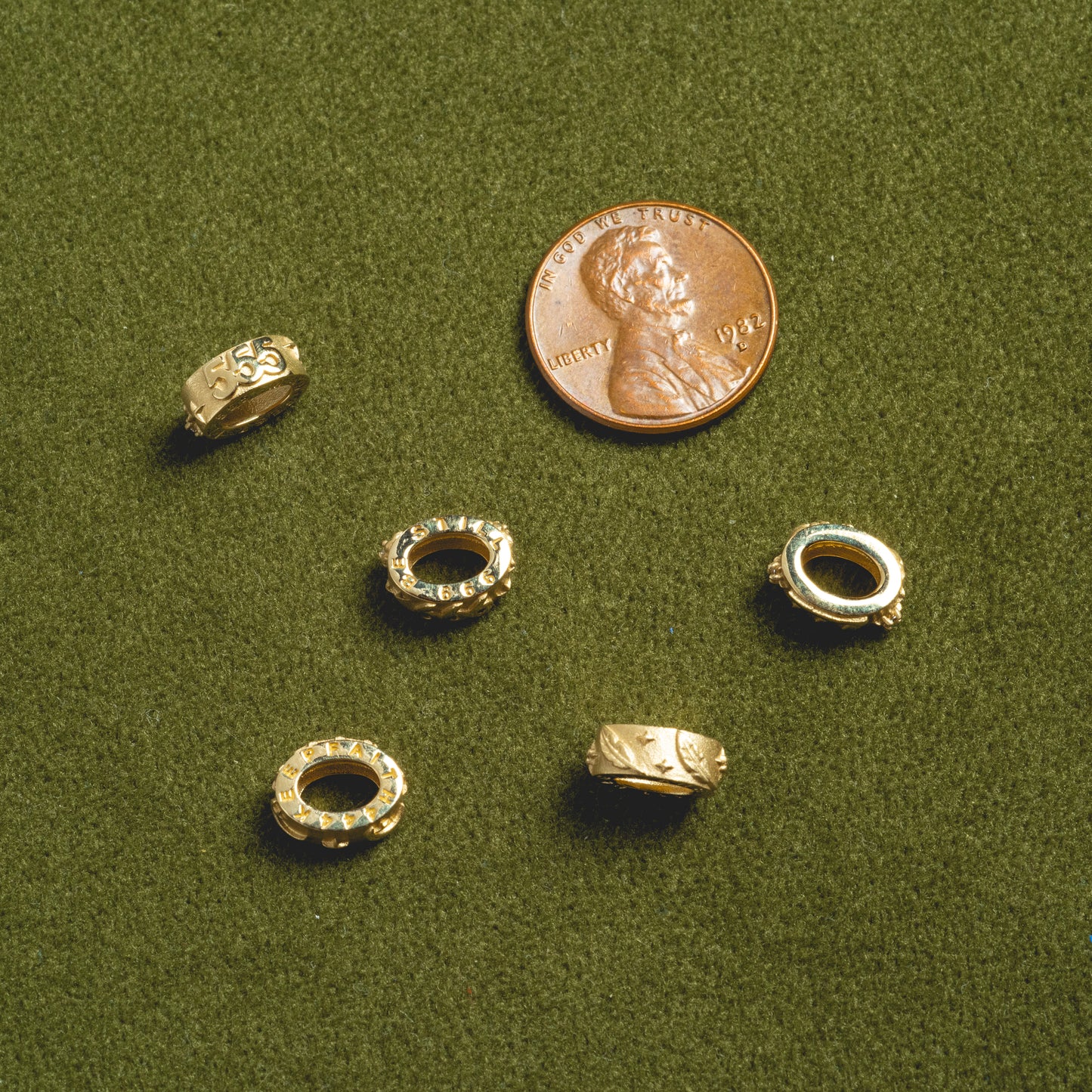 Five Solid Gold Angel Number Spacer Charms with engravings are displayed on a textured green surface next to a U.S. penny, highlighting their size for scale.