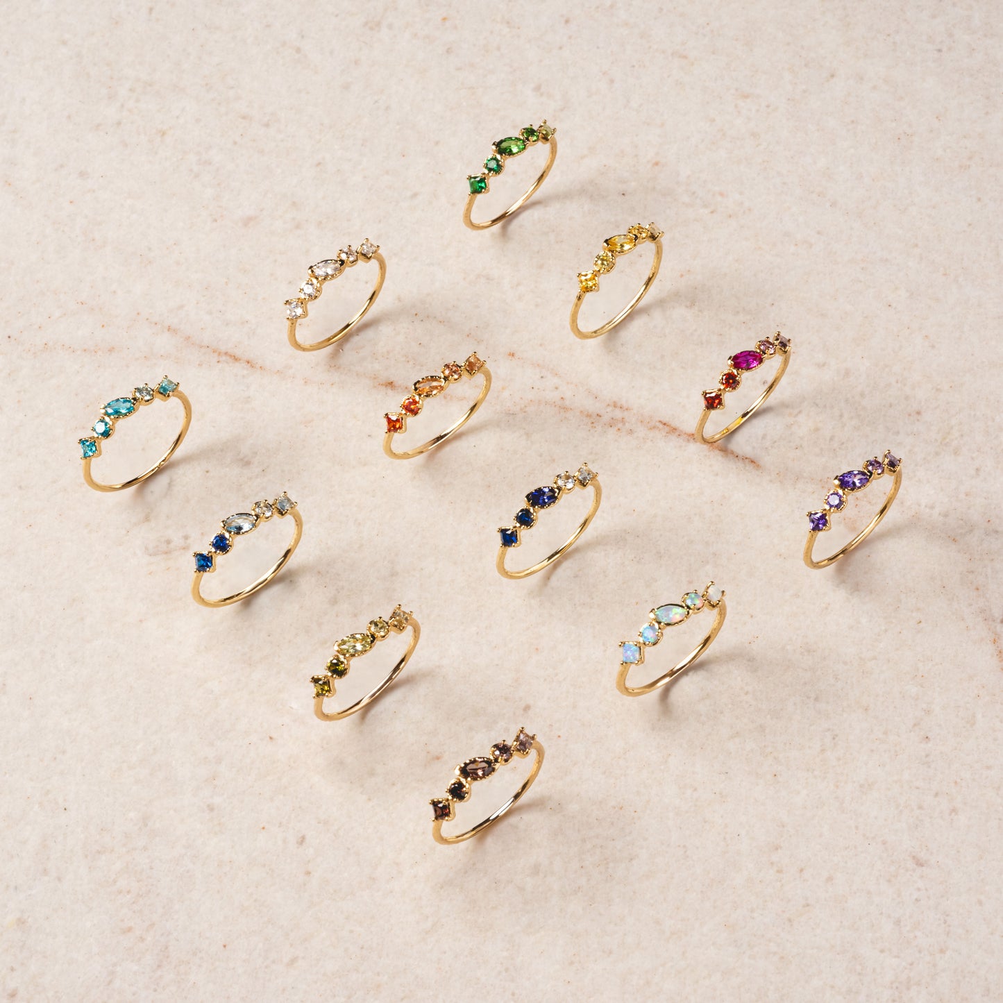 Twelve delicate 14k gold plated Shades of You Ombre Birthstone Rings with colorful gemstone clusters are arranged in neat rows on a light beige textured surface, each displaying a unique combination of gemstone colors.