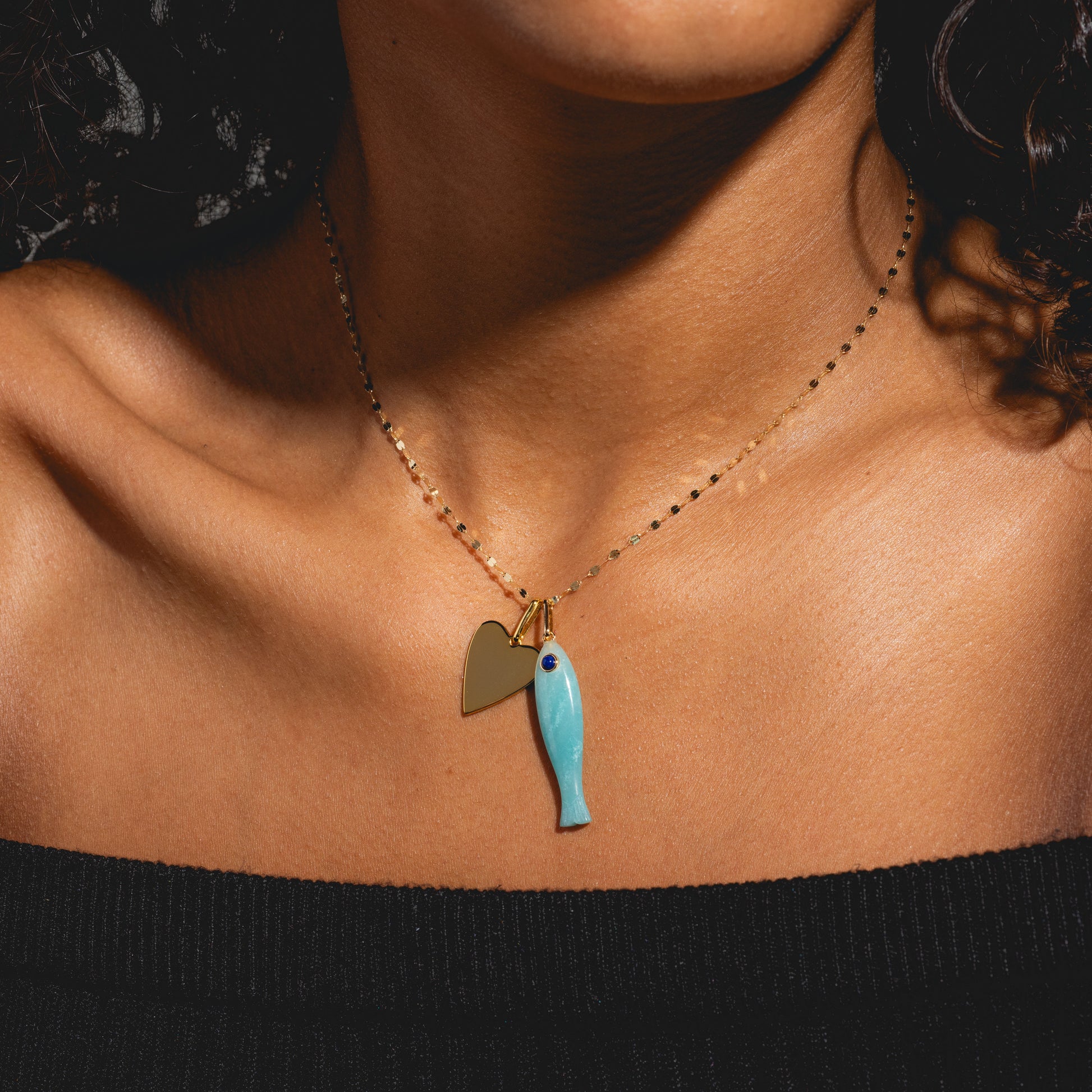 A close-up shows a person in a black off-shoulder top wearing Charms by Thatch: a gold necklace with personalized gold heart and blue fish charms, complementing their warm skin tone.