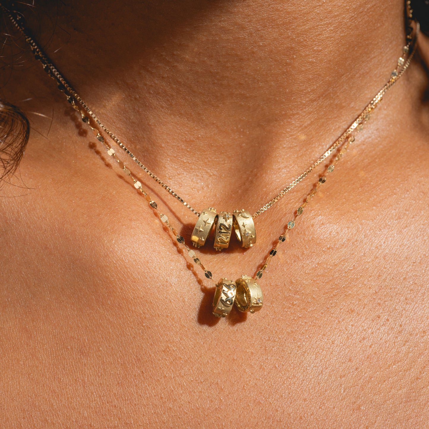 A close-up of a person’s neck wearing two layered gold necklaces with Solid Gold Angel Number Spacer Charms as pendants on delicate, beaded chains, glowing against sunlit, tan skin. A Local Eclectic Exclusive.