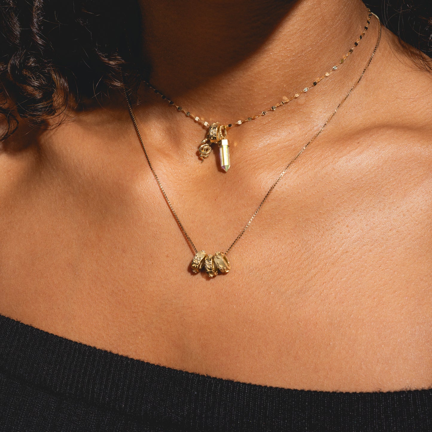 A close-up shows a person with warm brown skin wearing two gold necklaces—one featuring the Solid Gold Angel Number Spacer Charm and another with three textured beads—paired with a black off-the-shoulder top.