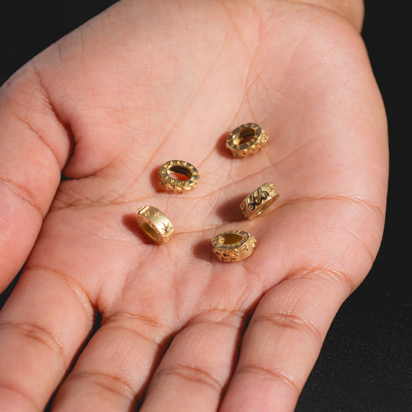A close-up of an open hand holding five small, round, gold-colored Solid Gold Angel Number Spacer Charms with engraved designs against a black background.