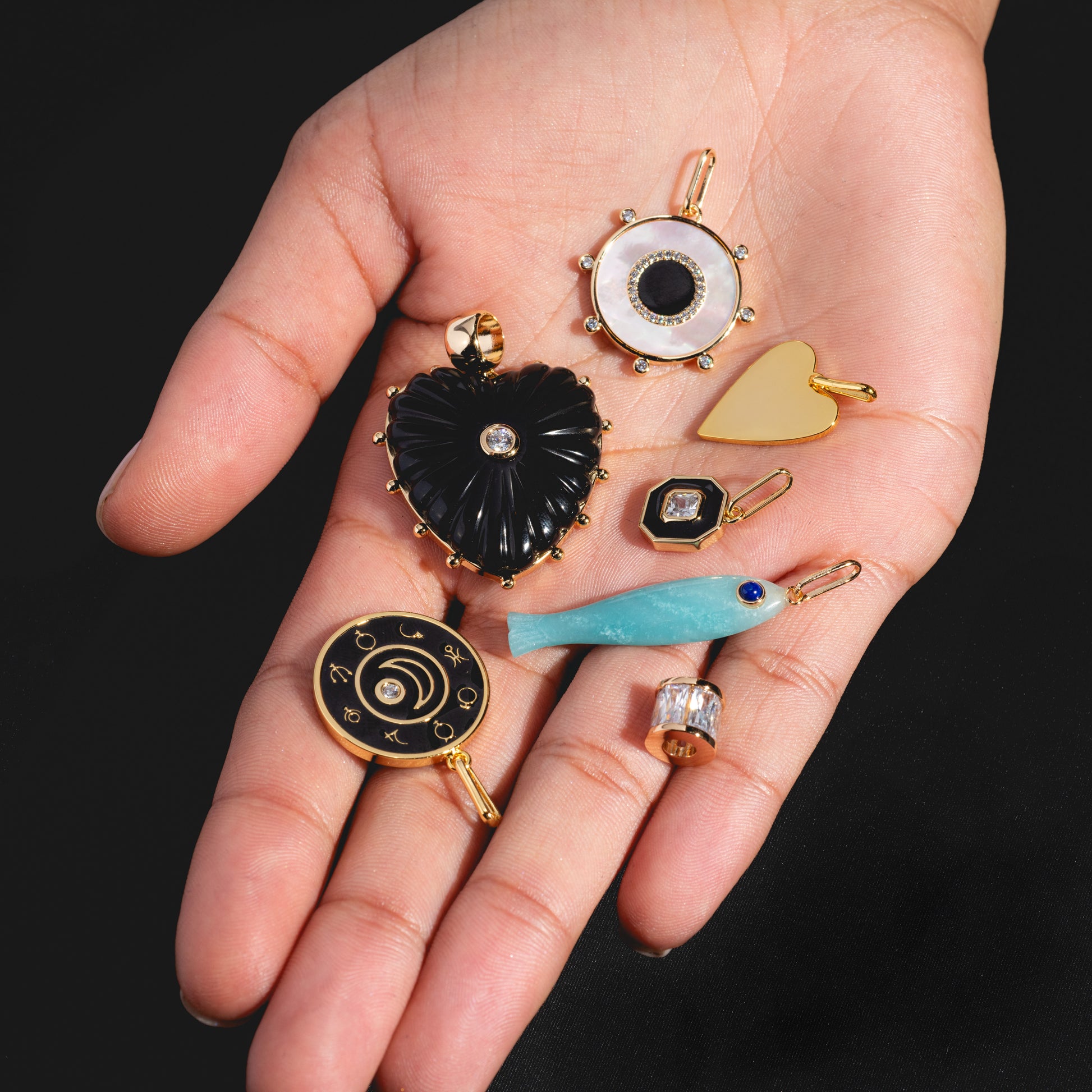 A hand displays Charms by Thatch—personalized necklace charms such as a black heart, blue fish, gold plated charm, mother of pearl charm, and an eye-shaped charm—all shown against a black background.