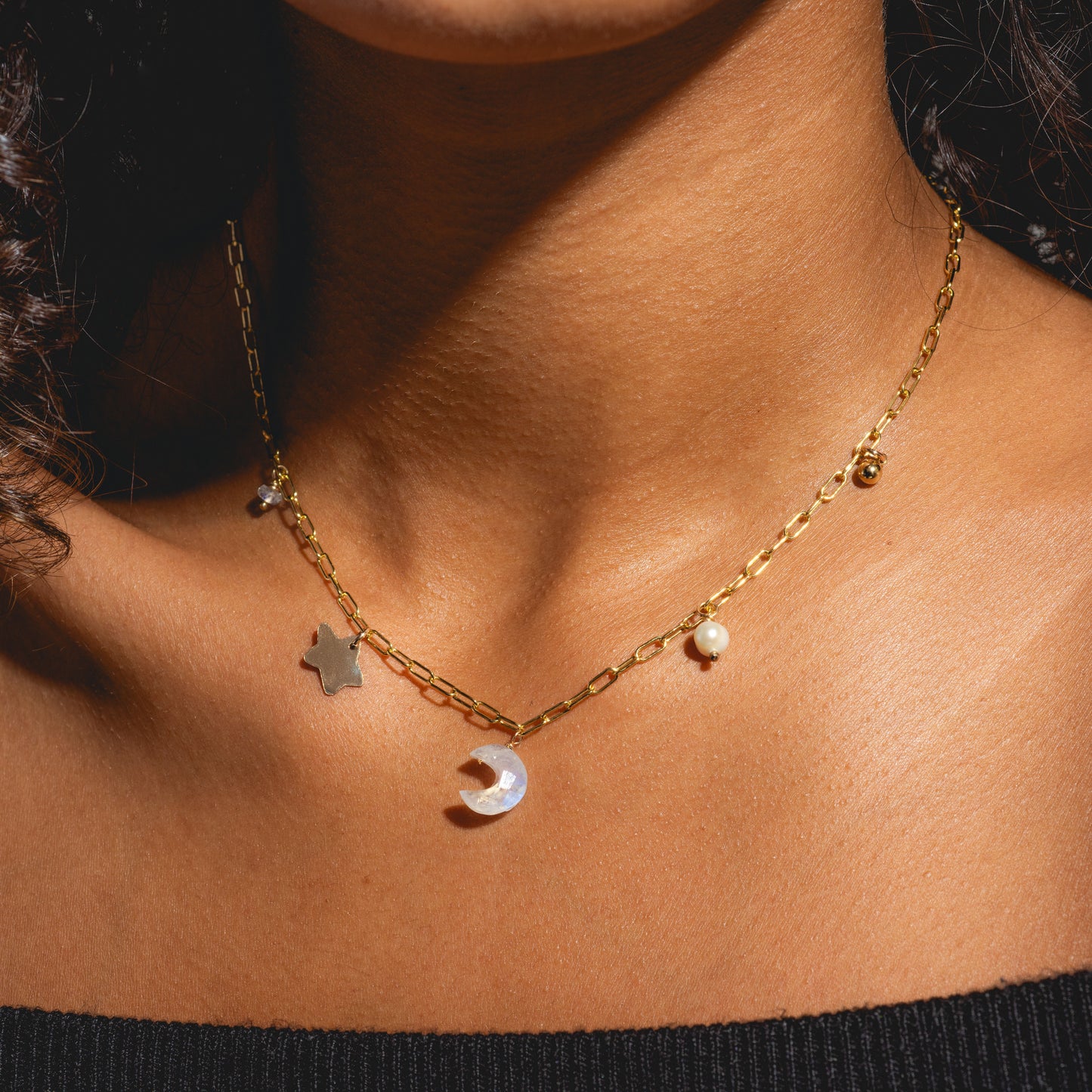 A close-up of a person wearing the Celine Charm Necklace Sample, a 14k gold filled chain with brass base star, crescent moon, and moonstone pearl charms, paired with a black off-the-shoulder top.
