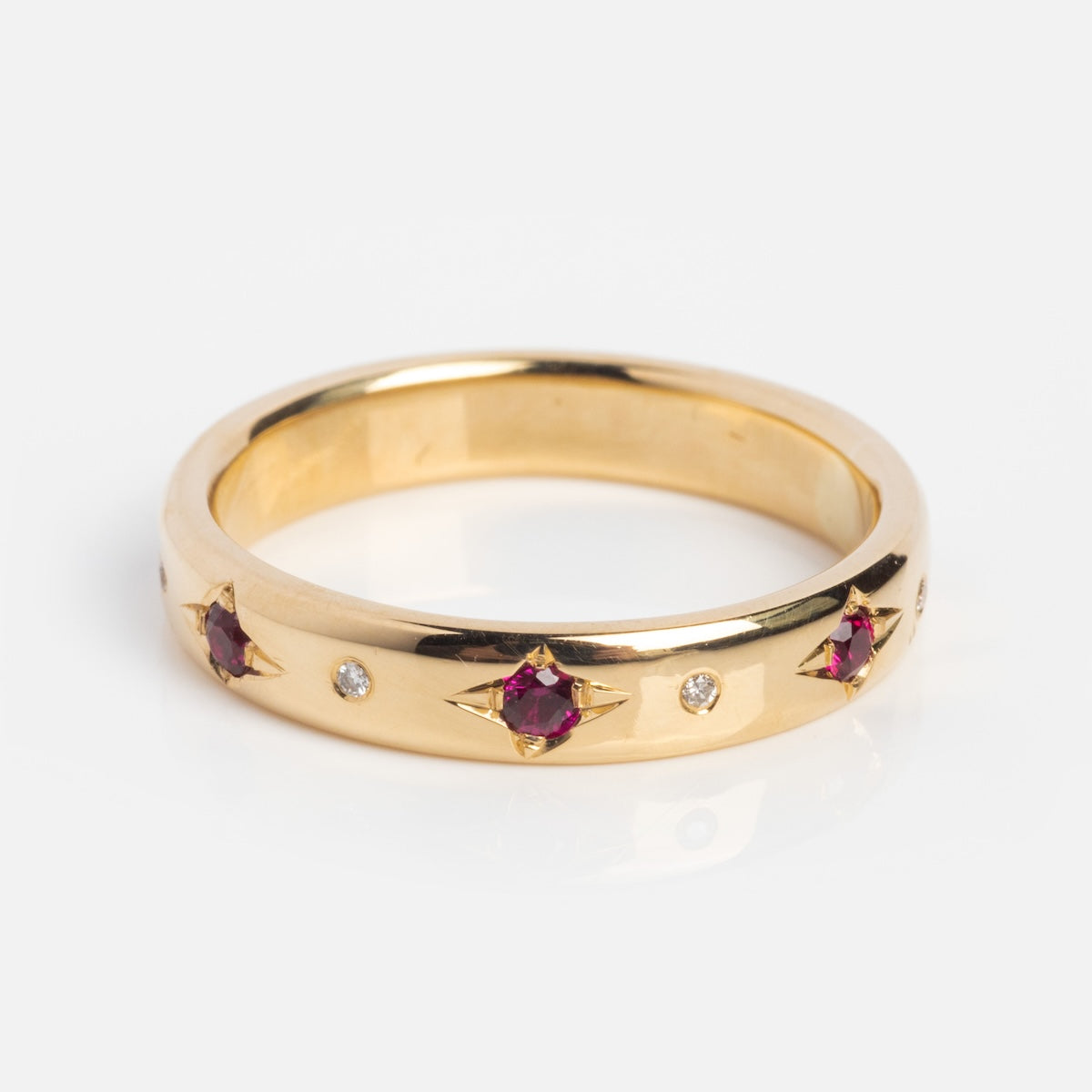 The Ruby and Diamond Eternity Band features 14k solid yellow gold with alternating round diamonds and star-shaped rubies, set around the band and displayed on a white background.