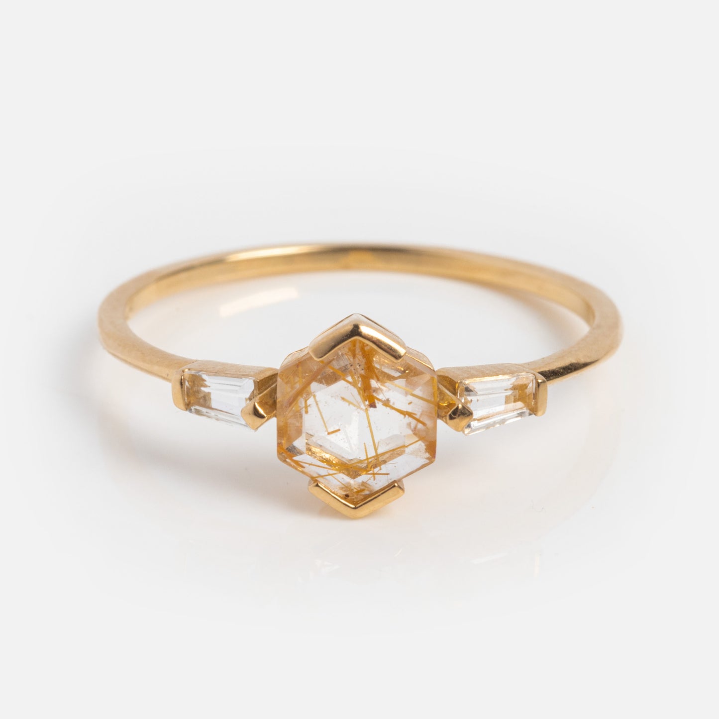 The Solid Gold Rutilated Quartz Illumination Ring features a central hexagonal rutilated quartz with golden inclusions, flanked by two clear rectangular stones on each side, set against a plain white background.