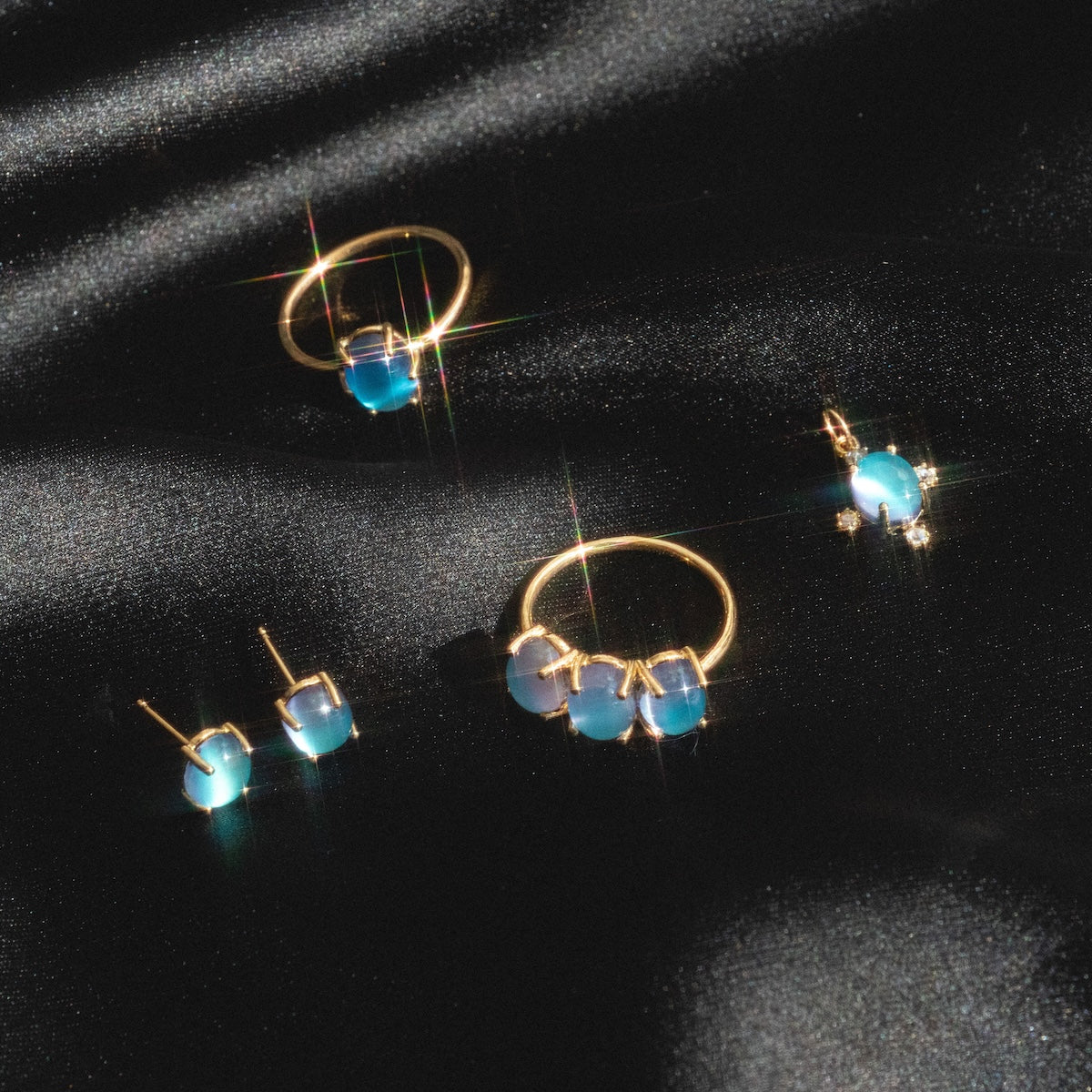 Gold jewelry with blue gemstones, including the Local Eclectic exclusive 14k Blue Crush Cat’s Eye Moonstone Baby Gumdrop Ring, plus hoop earrings, studs, and a pendant, displayed on black satin fabric with a sparkling effect.
