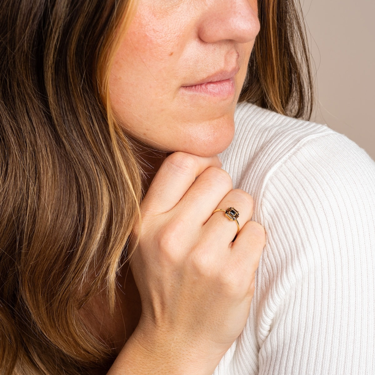 A woman with long brown hair rests her chin on her hand, wearing the 14k Vintage Inspired Art Deco Engagement Ring with a dark gemstone. Dressed in a white ribbed top, she gazes slightly to the side.