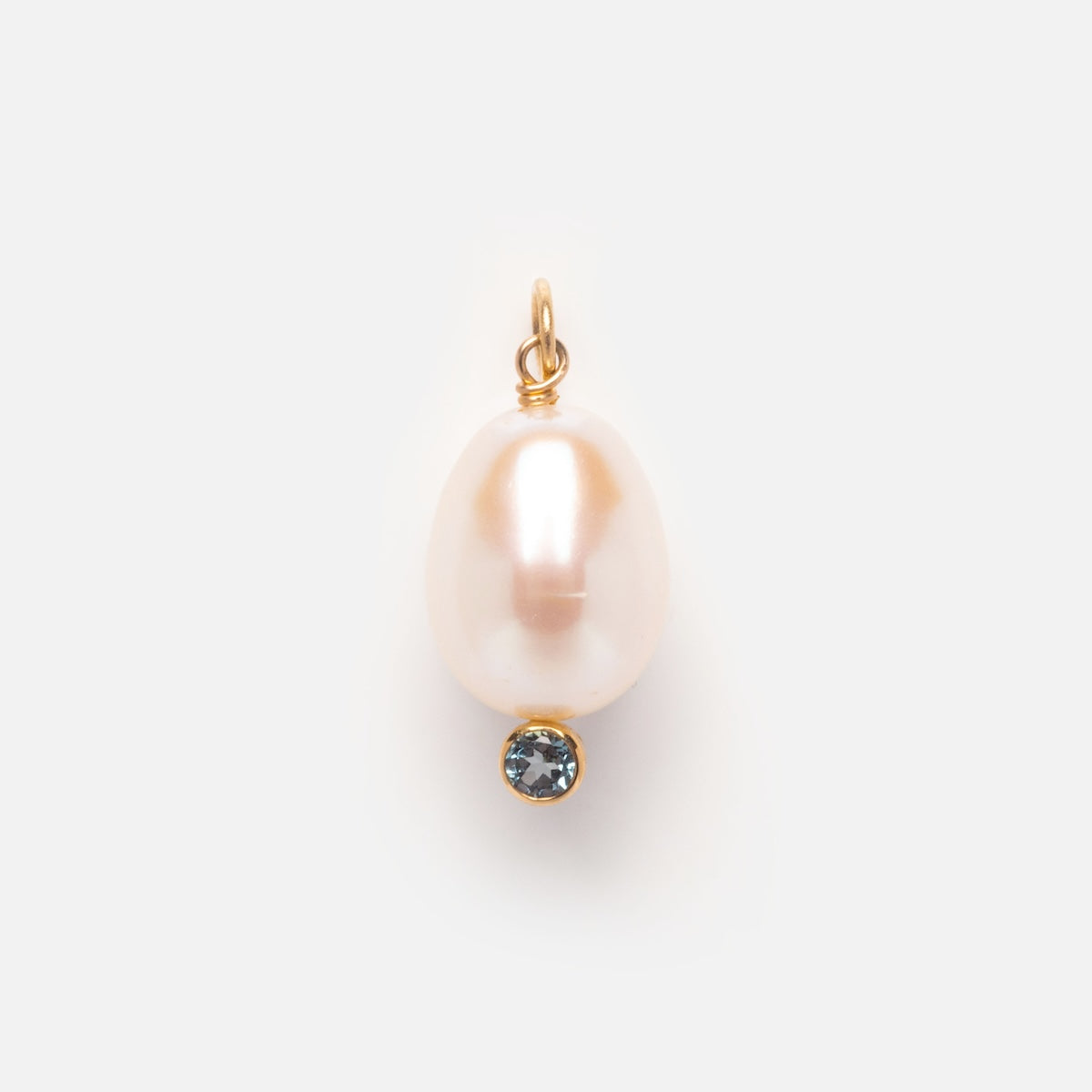 The Solid Gold Statement Charm features a single oval white pearl and a small round blue topaz set in 9k yellow gold, shown on a plain light gray background.