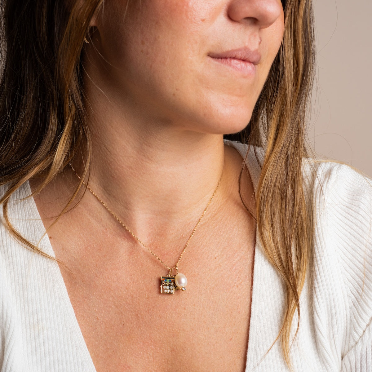 A woman with light brown hair wears a white ribbed top and a gold necklace featuring the Solid Gold Statement Charm, adorned with a pearl and blue topaz pendant, as she gazes elegantly to the side.