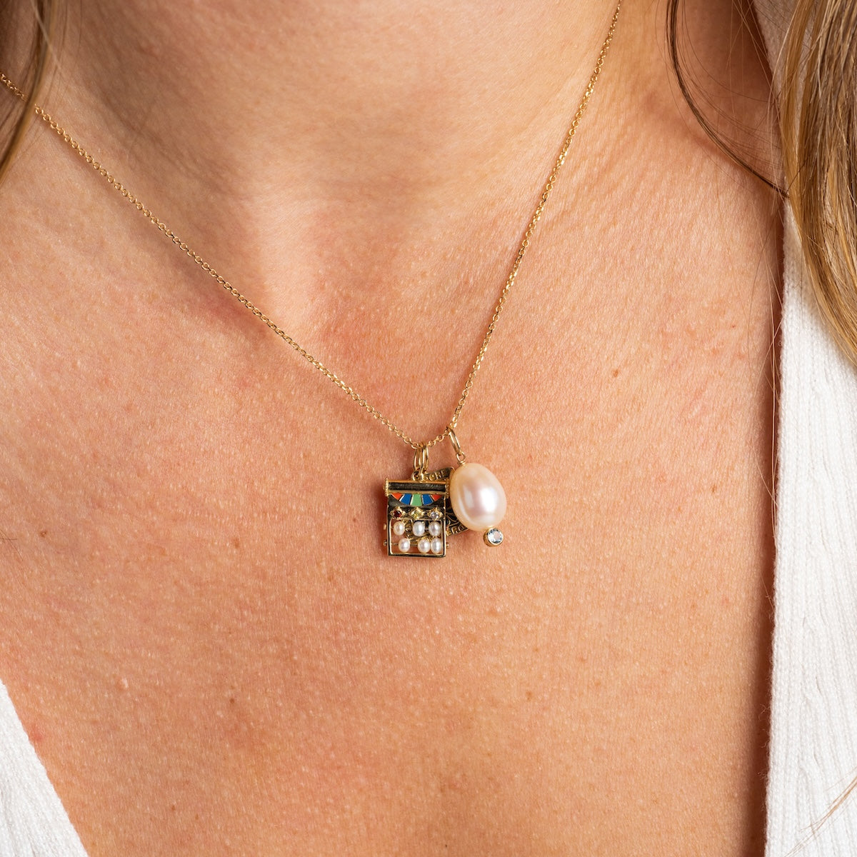 A close-up of the Solid Gold Statement Charm necklace, featuring a rectangular pendant with pearls and blue topaz plus a large pearl, styled above a white V-neck top.