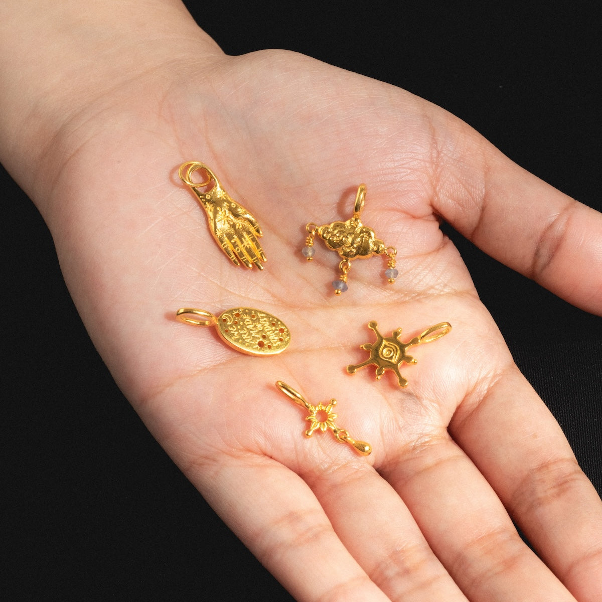A hand displays five unique gold vermeil charms from Charms by Hunt of Hounds: a hand, a crab, an oval with patterns, a sunburst, and a circular charm with an eye, all set against a black background.