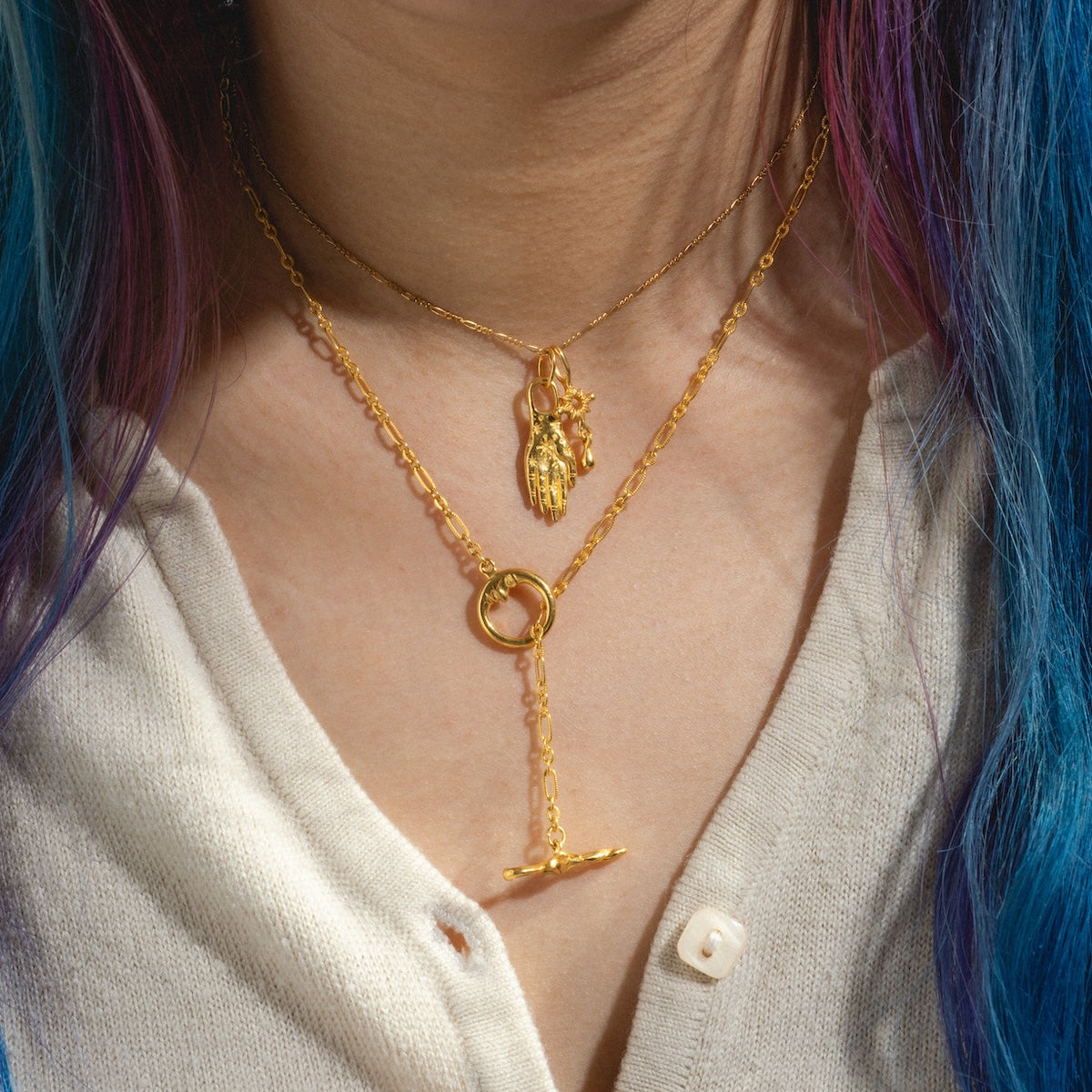 A person with purple and blue hair wears two layered Charms by Hunt of Hounds gold vermeil necklaces—a hand charm pendant and a toggle clasp with round ring—over a cream buttoned shirt.