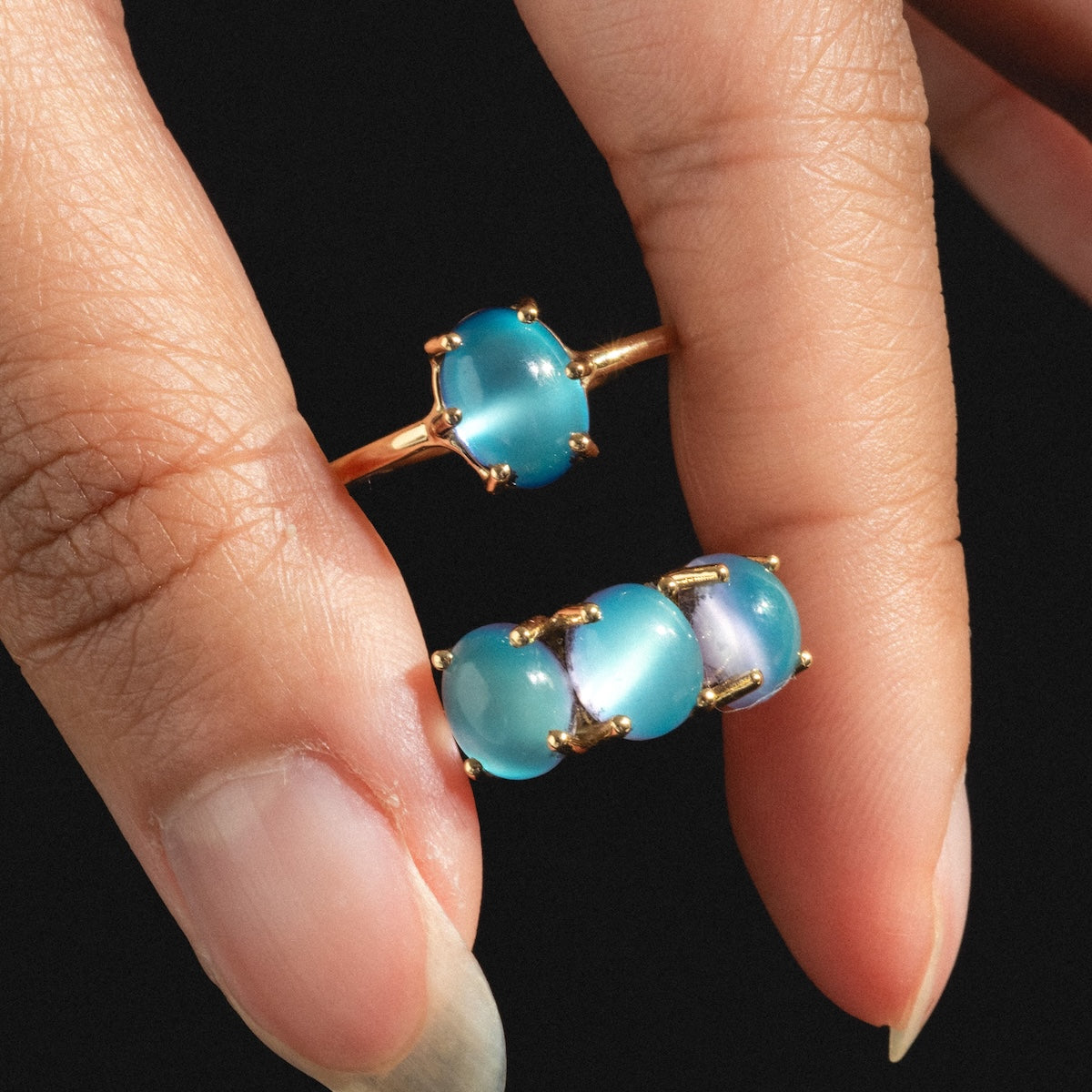A hand with long nails wears the Local Eclectic exclusive 14k Blue Crush Cats Eye Moonstone Baby Gumdrop Ring and holds another matching ring, both shown against a black background.