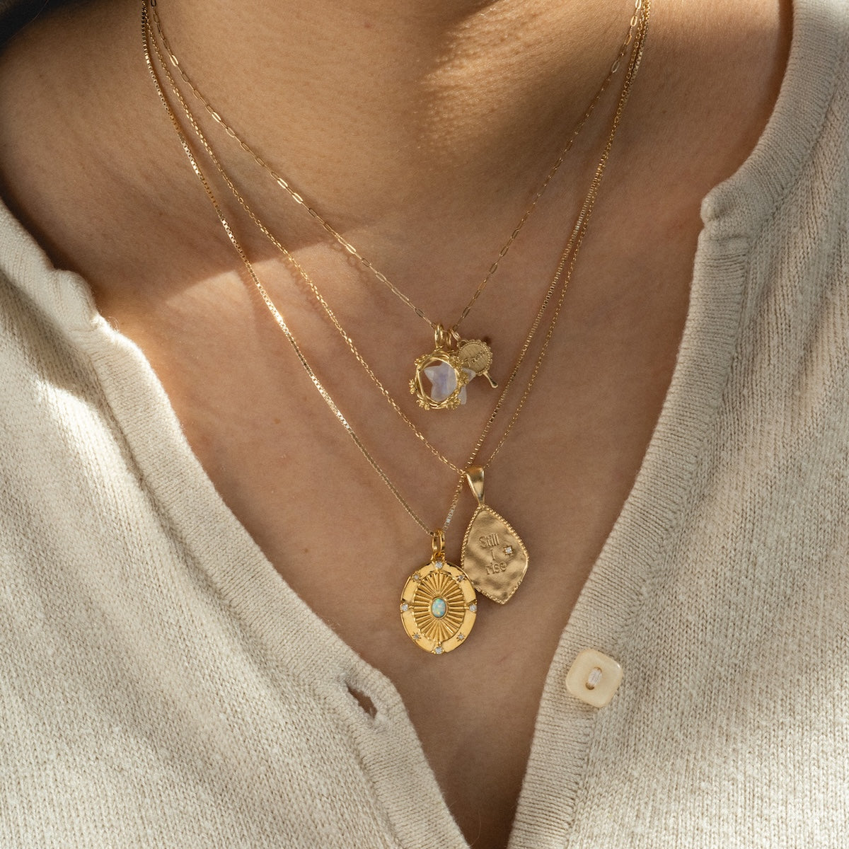 A person in a cream buttoned shirt showcases three layered gold necklaces from the Limited Edition Womens History Month Designer Charms, each with a unique pendant: a small gemstone, an oval medallion, and a rectangular engraved charm.
