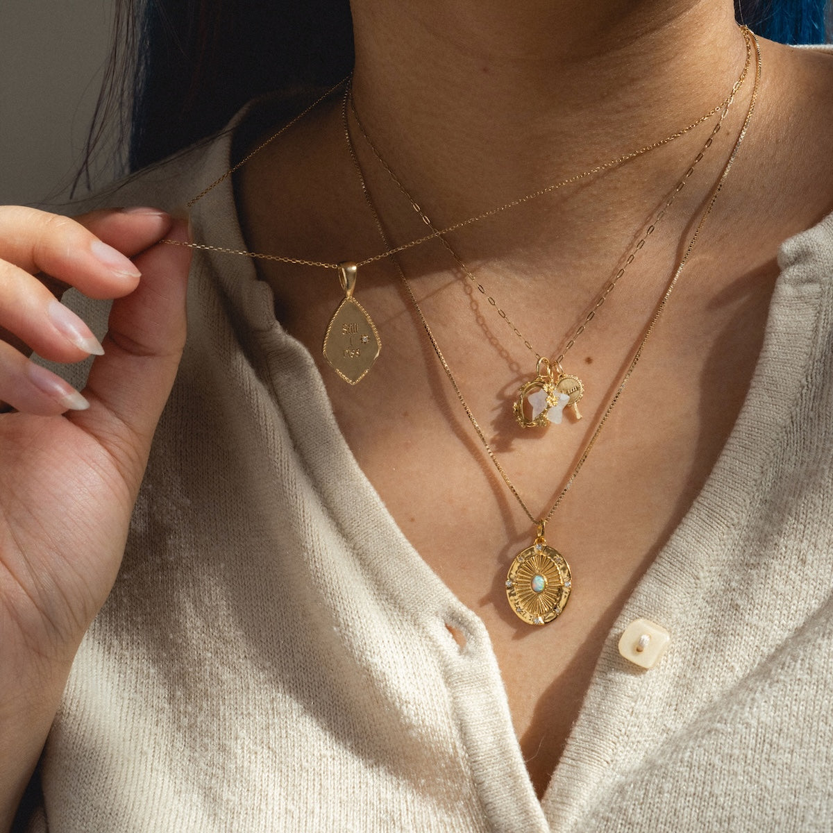 A person wears a beige buttoned top and three layered 14k gold plated Charms by Joy Dravecky necklaces, featuring a leaf pendant, a blue-centered circular charm, and a cluster of small stones.