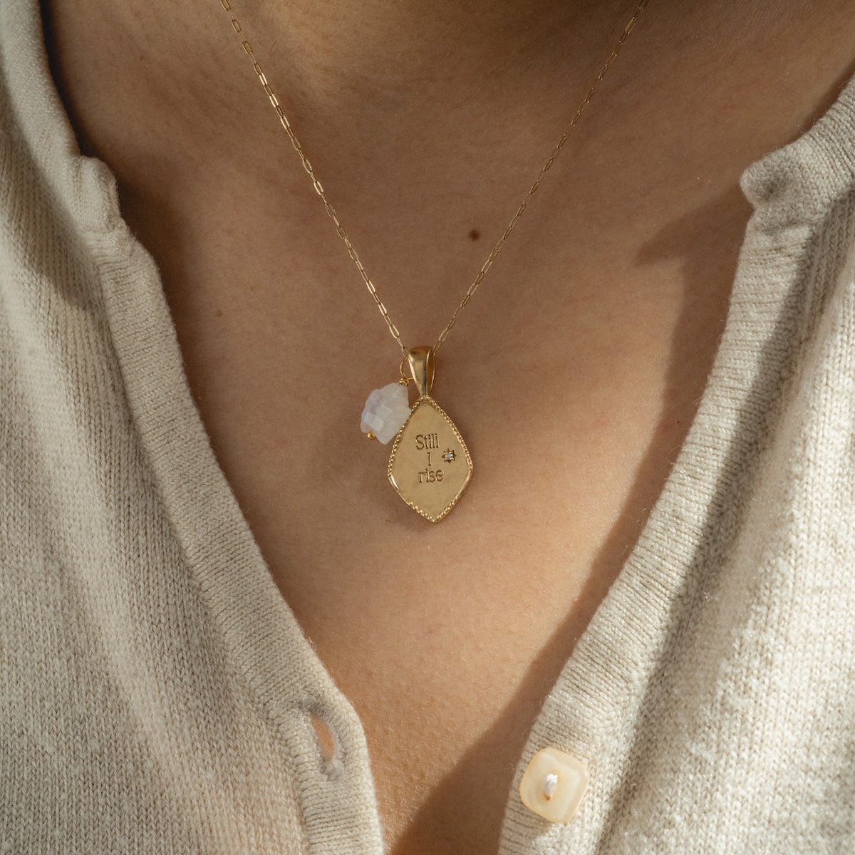 A close-up of a person wearing the Charms by Joy Dravecky 14k gold plated necklace with an engraved smil* rts pendant and a small white stone, styled over a beige, buttoned-up knit top.