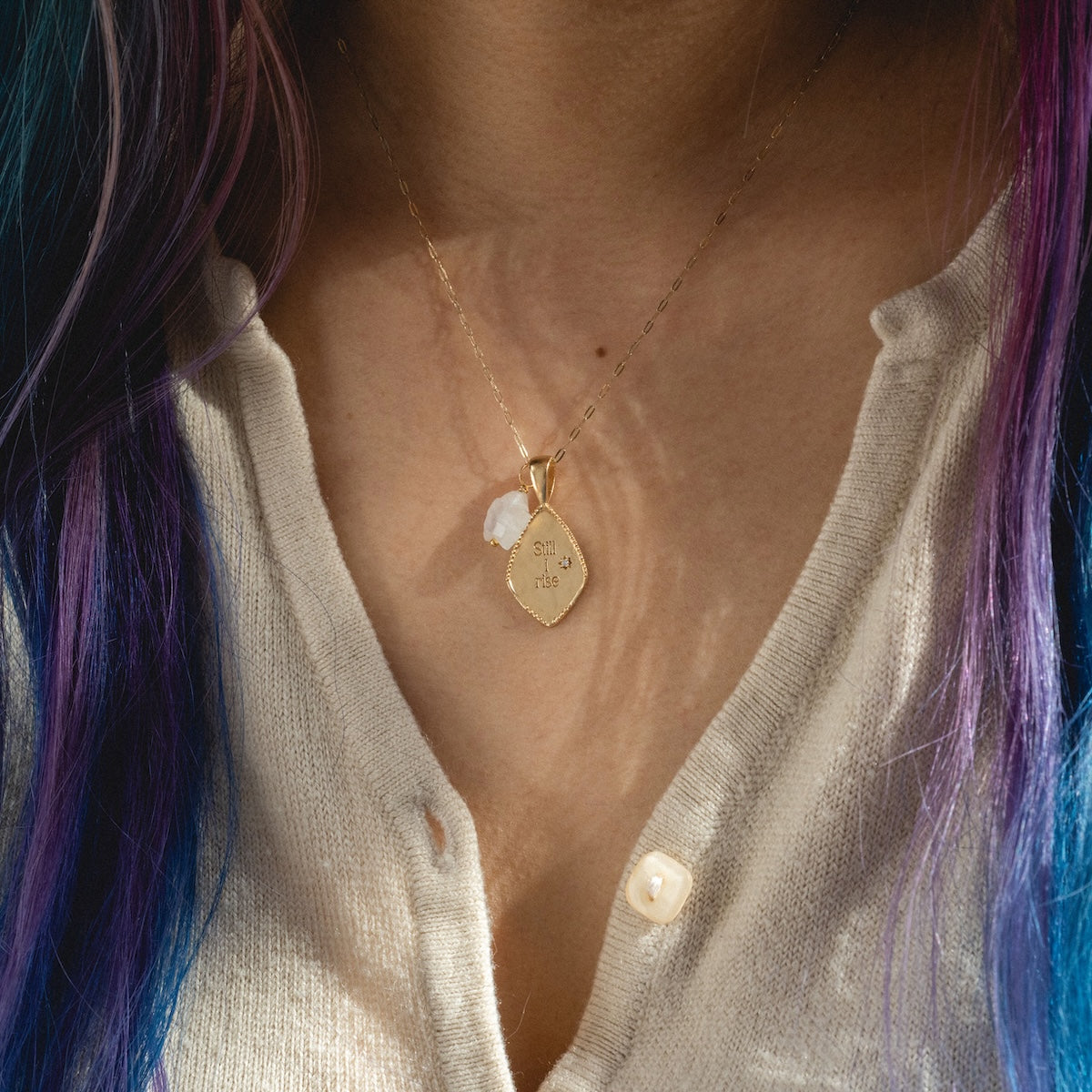 A person with long, wavy, purple and blue hair wears a beige buttoned cardigan and Charms by Joy Dravecky—a 14k gold plated necklace featuring oui and rêve charms plus a small white charm.