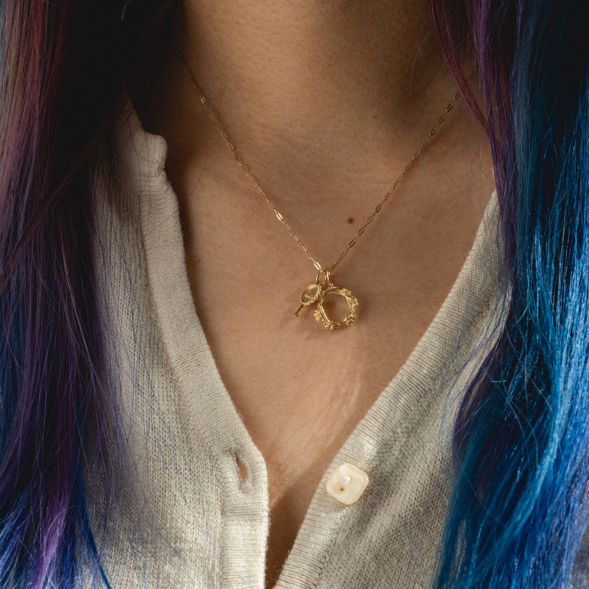 A person with long blue and purple hair wears a cream-colored buttoned shirt and a delicate Charlie and Marcelle 14k Littlest Charms necklace, featuring gold charms including a wreath-shaped circular pendant with a small bow.