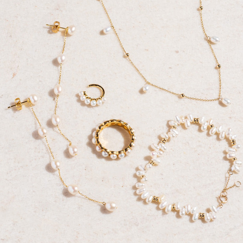 A flat lay of gold jewelry with pearls, featuring two necklaces, a bracelet, Delicate Pearl Drop Earrings, a single hoop earring, and a gold ring—all on a light beige surface. A Local Eclectic exclusive in 14k yellow gold plating.