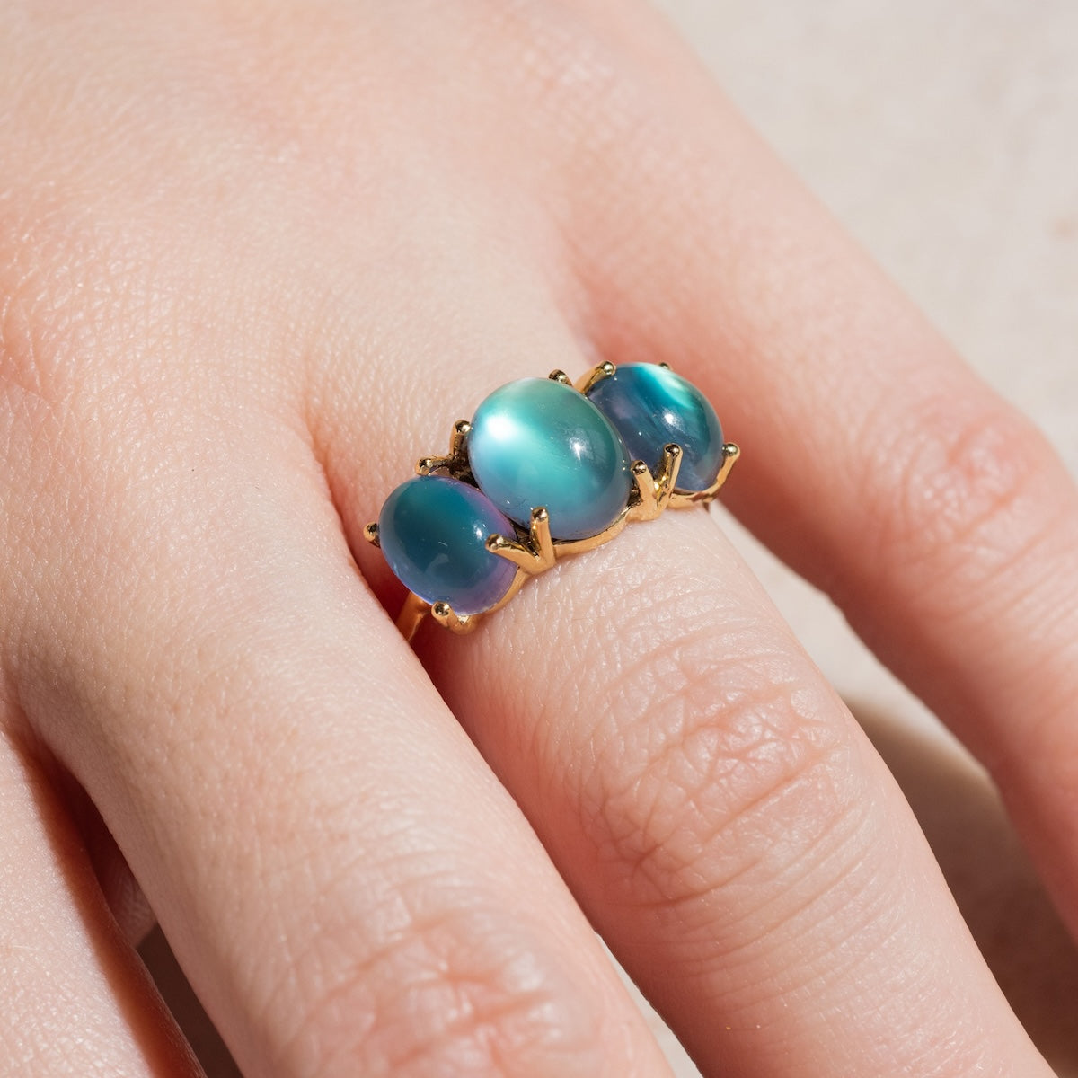 A hand with a light complexion wears the Sweetest Gumdrop Ring, crafted in yellow gold and set with three oval iridescent blue-green gemstones. Each stone is secured by four prongs, and the ring is worn on the middle finger.