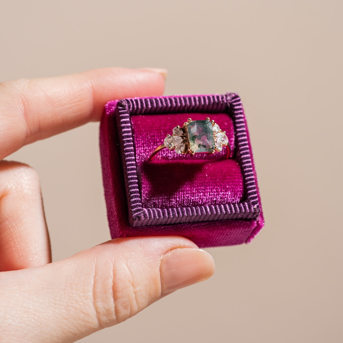 A hand holds the Gemstone Empress Ring, a gold statement piece featuring a rectangular multicolored gemstone between two clear stones, displayed in a fuchsia velvet box against a beige background.