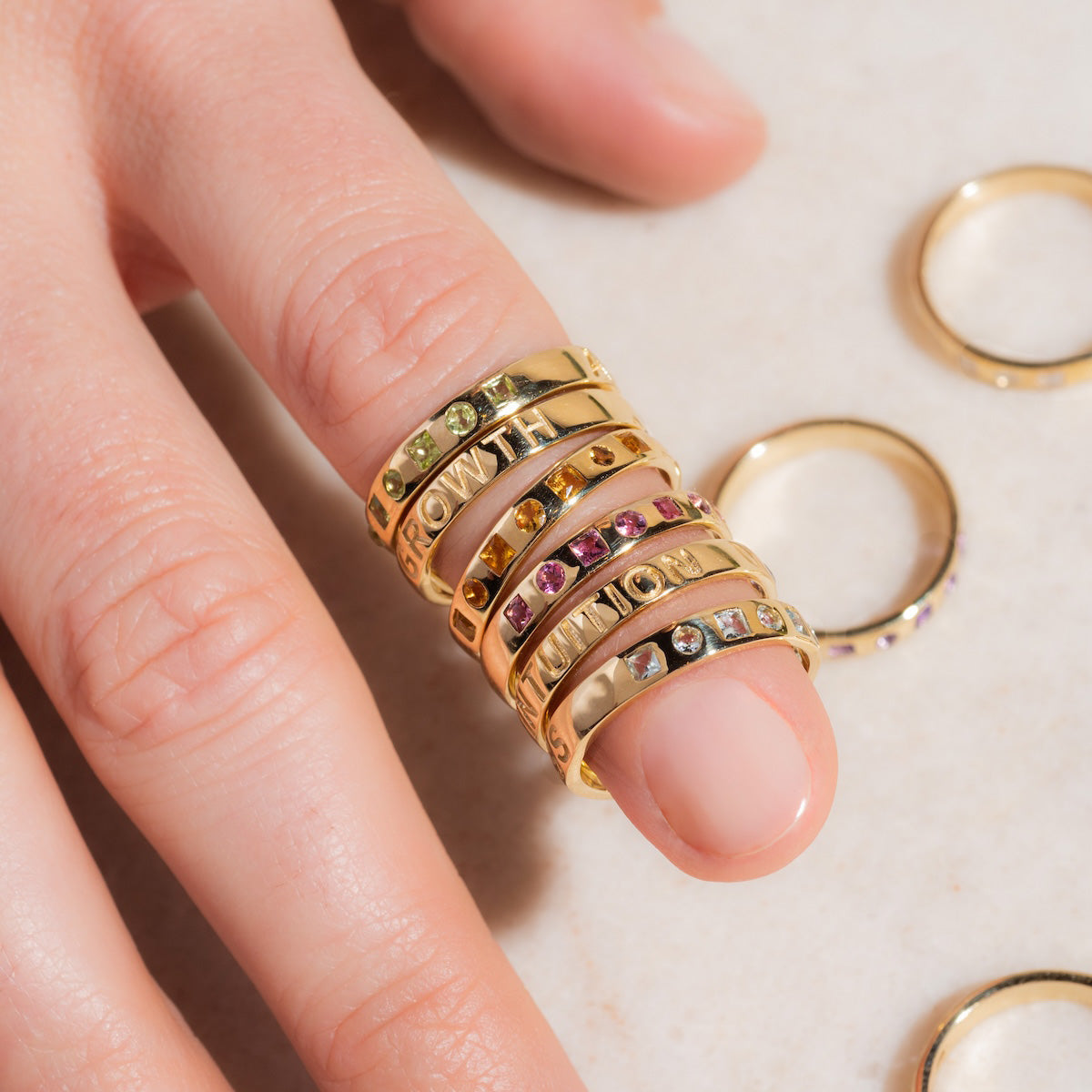 A hand displays Solid Gold Within You Birthstone Band (sample size 7) rings in 9k yellow gold, some set with natural stones and engraved words, resting on a light surface. Extra 9k gold bands (3.1 mm wide) are scattered nearby.