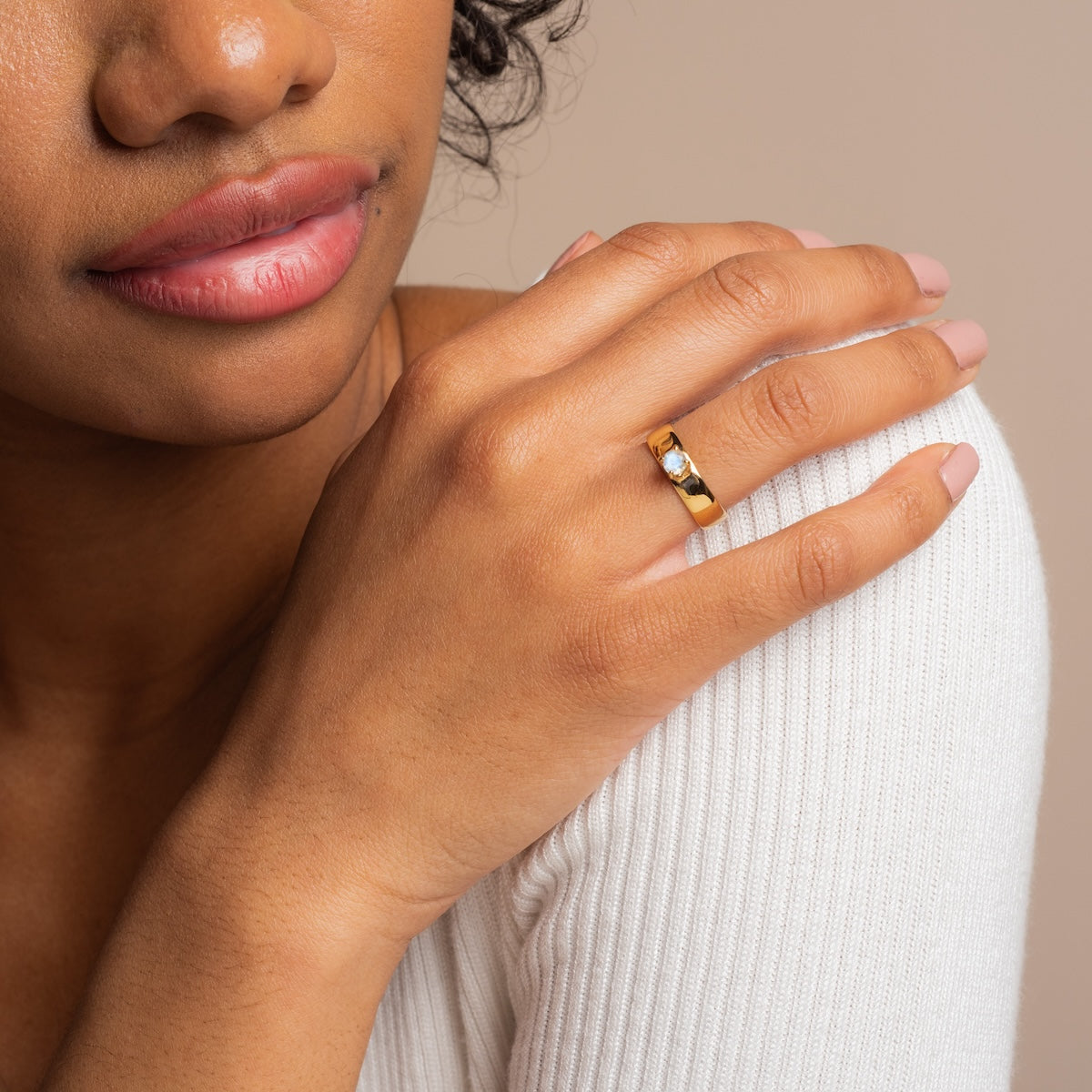 A woman in a white ribbed top rests her hand on her shoulder, showcasing the Lucinda Moonstone Ring Sample Size 6 in 14k yellow gold plating with a 5mm band. Her nails are neatly manicured with light pink polish.