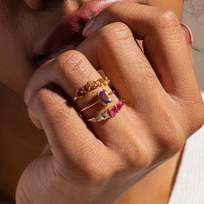A close-up of a hand wearing four Solid Gold Essential Baguette Birthstone Rings with vibrant gemstones, held near the lips. The softly blurred background and bright lighting highlight the rings’ colorful beauty.