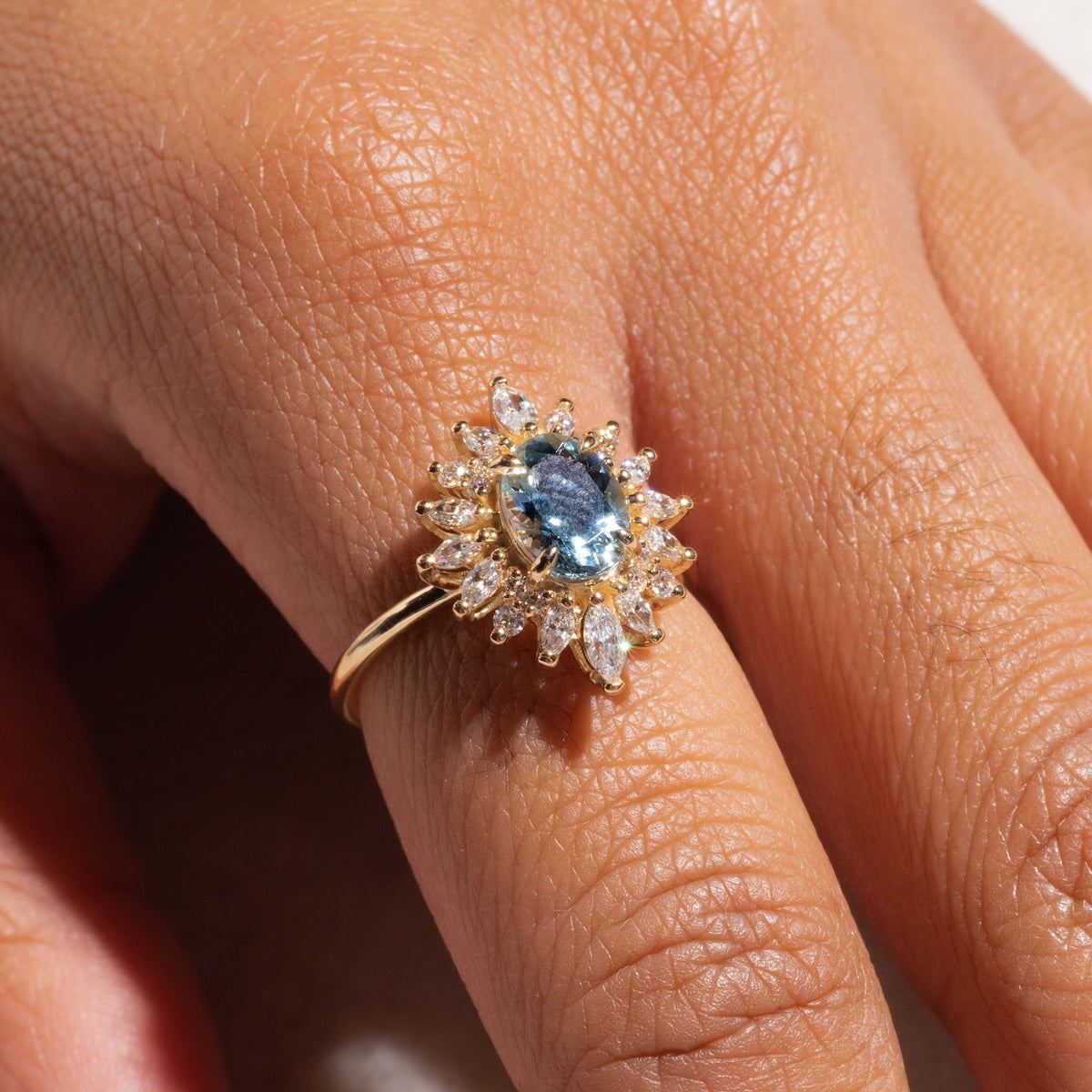 A close-up of a hand wearing the Lana Ring with Aquamarine & CZ, showcasing an oval blue aquamarine gemstone encircled by sparkling cubic zirconia in a sunburst halo pattern, all set in 14k solid gold.