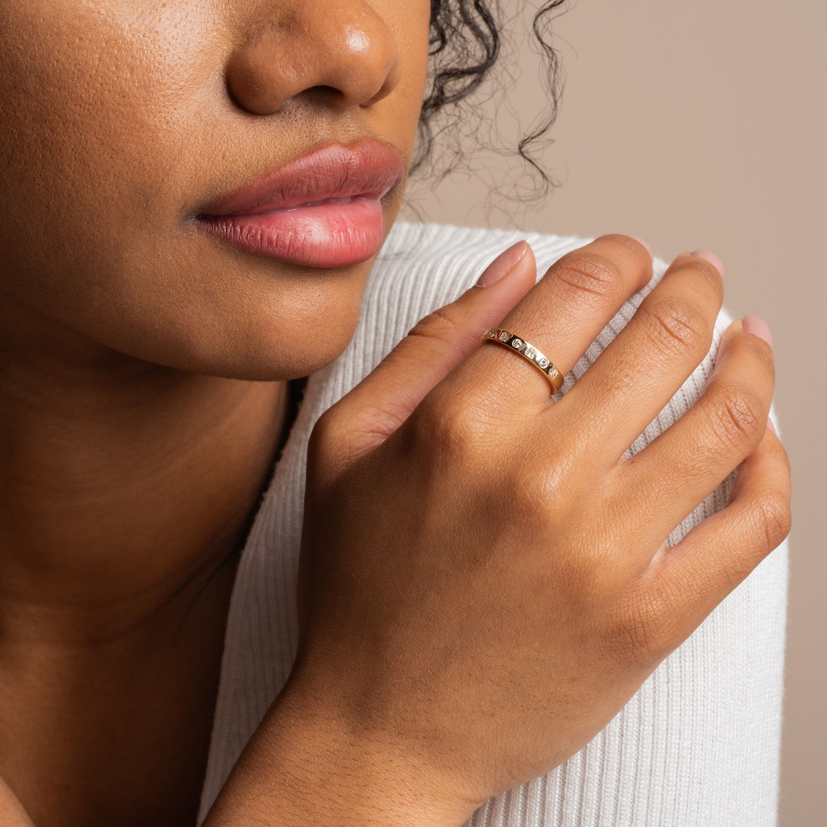 A woman in a white ribbed top gently touches her face, highlighting the Solid Gold 2024 April Capsule Diamond Statement Band (Sample Size 7) on her finger. The focus is on her hand, lips, and the sparkling lab diamond ring.