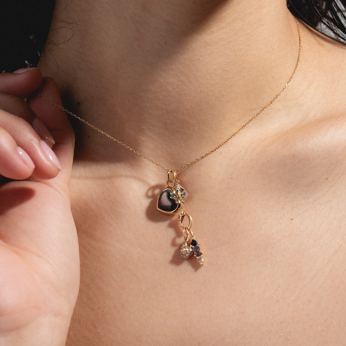 A close-up of a person wearing the Charms by Elizabeth Stone necklace, 14k gold plated with three charms—a black heart, a small crystal, and a cluster of crystals—while gently touching it near their collarbone.