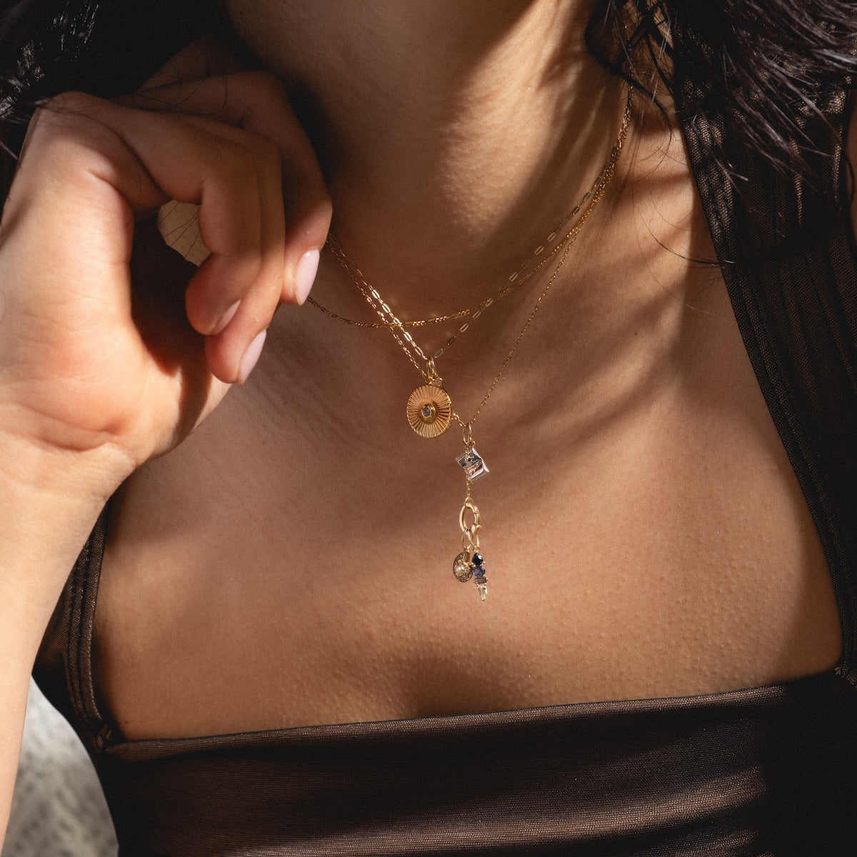 A close-up of a woman’s neck adorned with the Solid Gold Chain for Charms, featuring layered gold pieces like a spiral disc and small charms. Her hand rests by her face, and she wears a dark, sheer top.