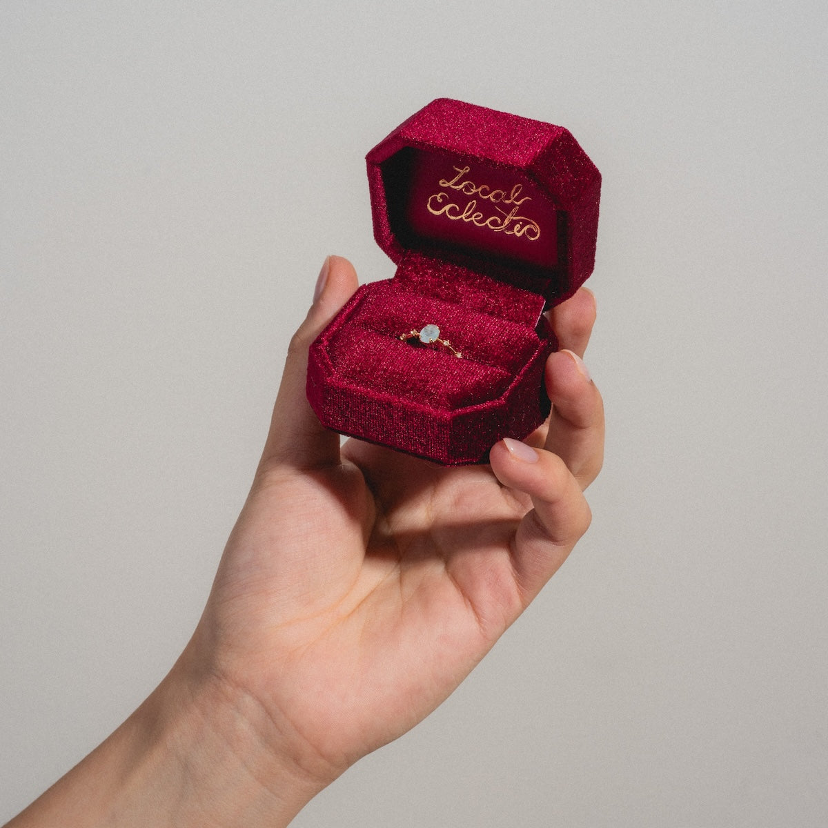 A hand holds an open Fancy Velvet Ring Box in rich red velvet, showing a delicate gold ring with a small gemstone. Inside the lid, Local Eclectic is written in elegant gold script.