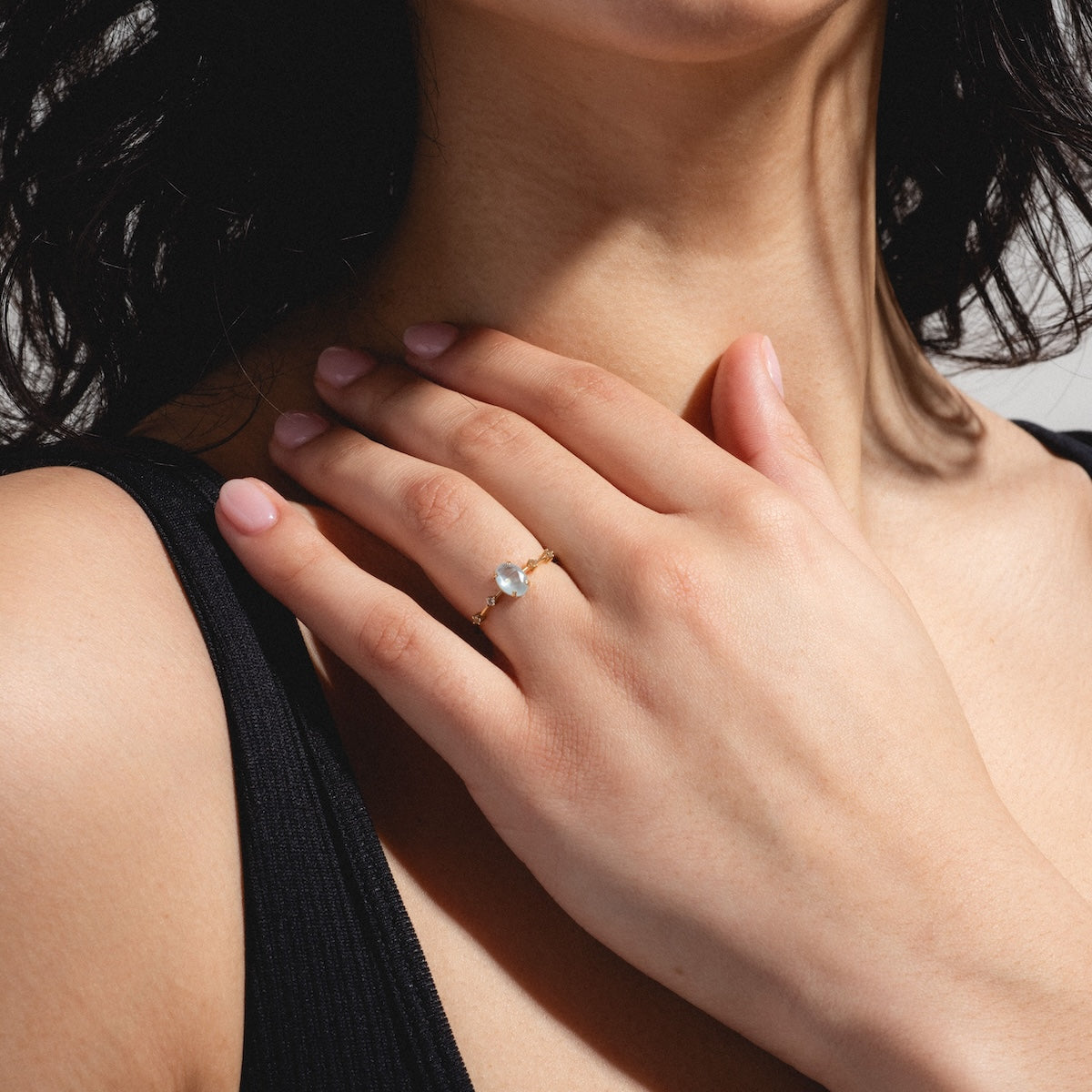 A woman with wavy dark hair displays the Solid Gold 2025 March Capsule Classic Aquamarine Statement Ring on her left hand. She wears a black sleeveless top and has natural-colored manicured nails—a chic March accessory.