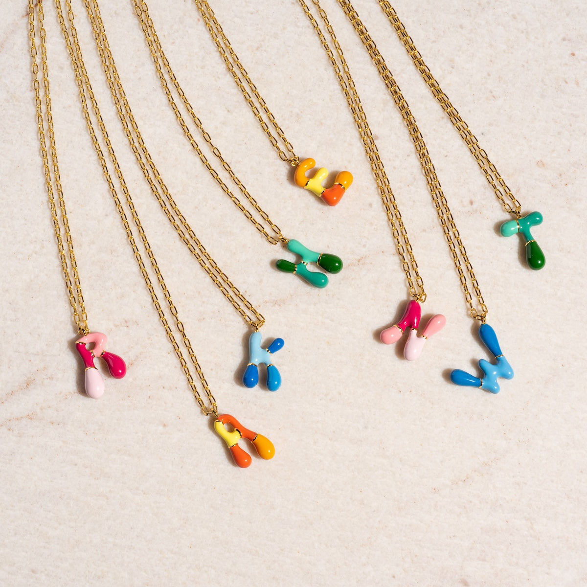 The Blue Bubble Letter Necklace Sample features a brass chain with 14k gold plating and a colorful, abstract resin letter pendant. Each unique pendant displays bright colors and is shown arranged on a light marble surface.