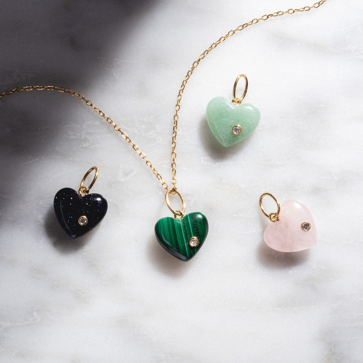 The Solid Gold Full Heart Crystal Charm is a necklace with a green crystal heart pendant and three gemstone-accented heart charms in black, green, and pink, beautifully displayed on a light marble surface.
